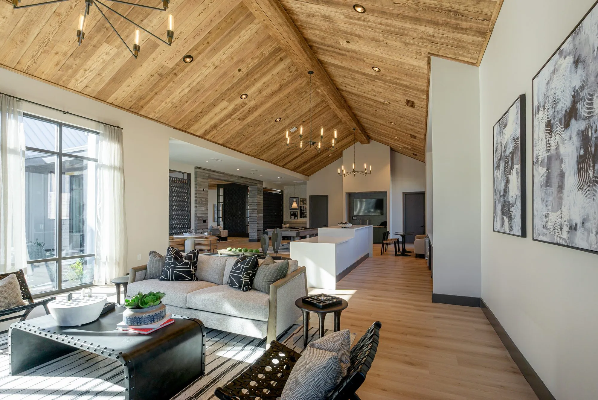 Multifamily amenity clubroom under a plank vaulted ceiling, grey sofa and riveted coffee table toward a white island