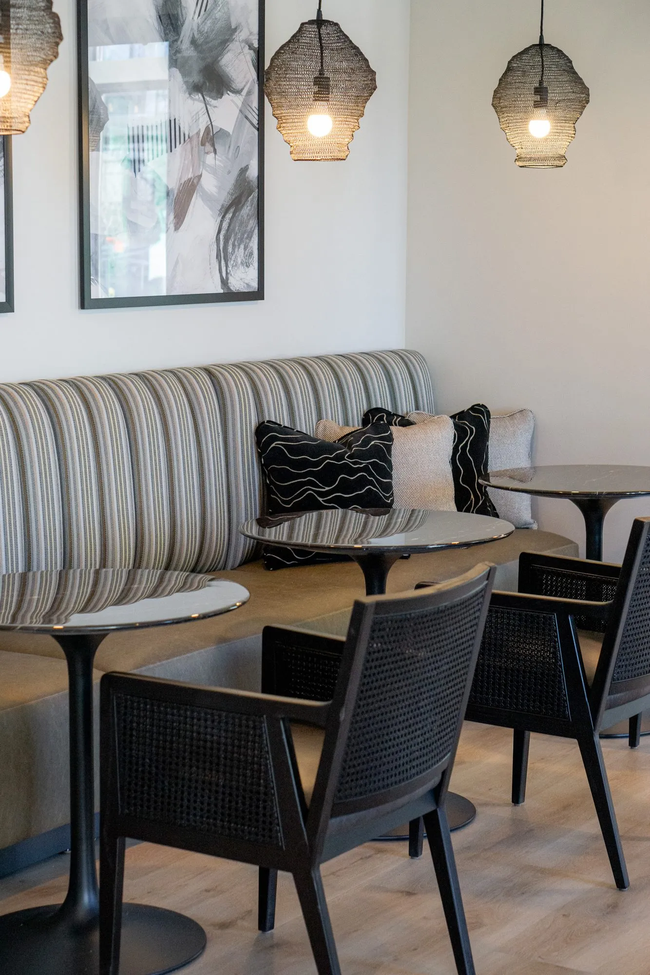 Multifamily amenity banquette with black cane chairs at round tables under wire mesh pendants, framed botanical prints above