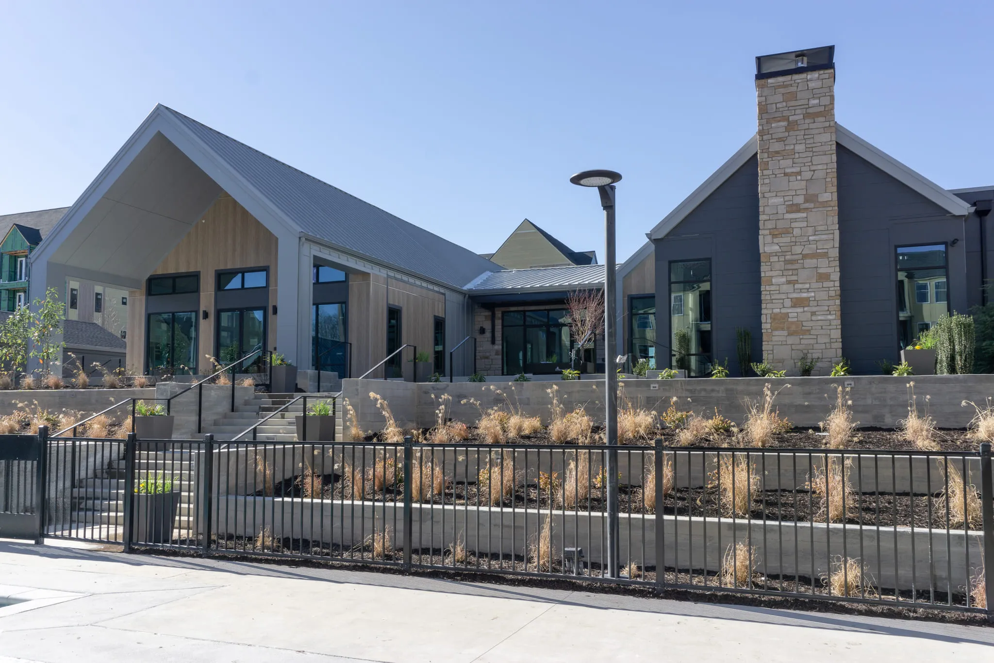 Multifamily community exterior: clubhouse with a gabled metal roof and stone chimney above a fenced planting bed