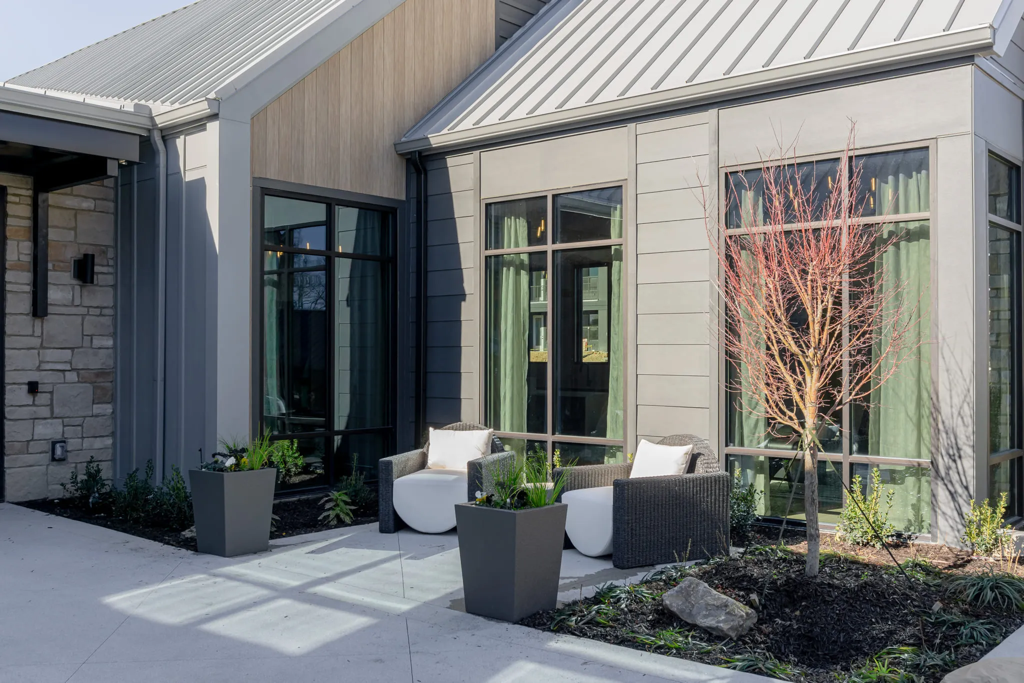 Multifamily amenity courtyard with woven lounge chairs on white cushions and planted beds against the clubhouse glazing