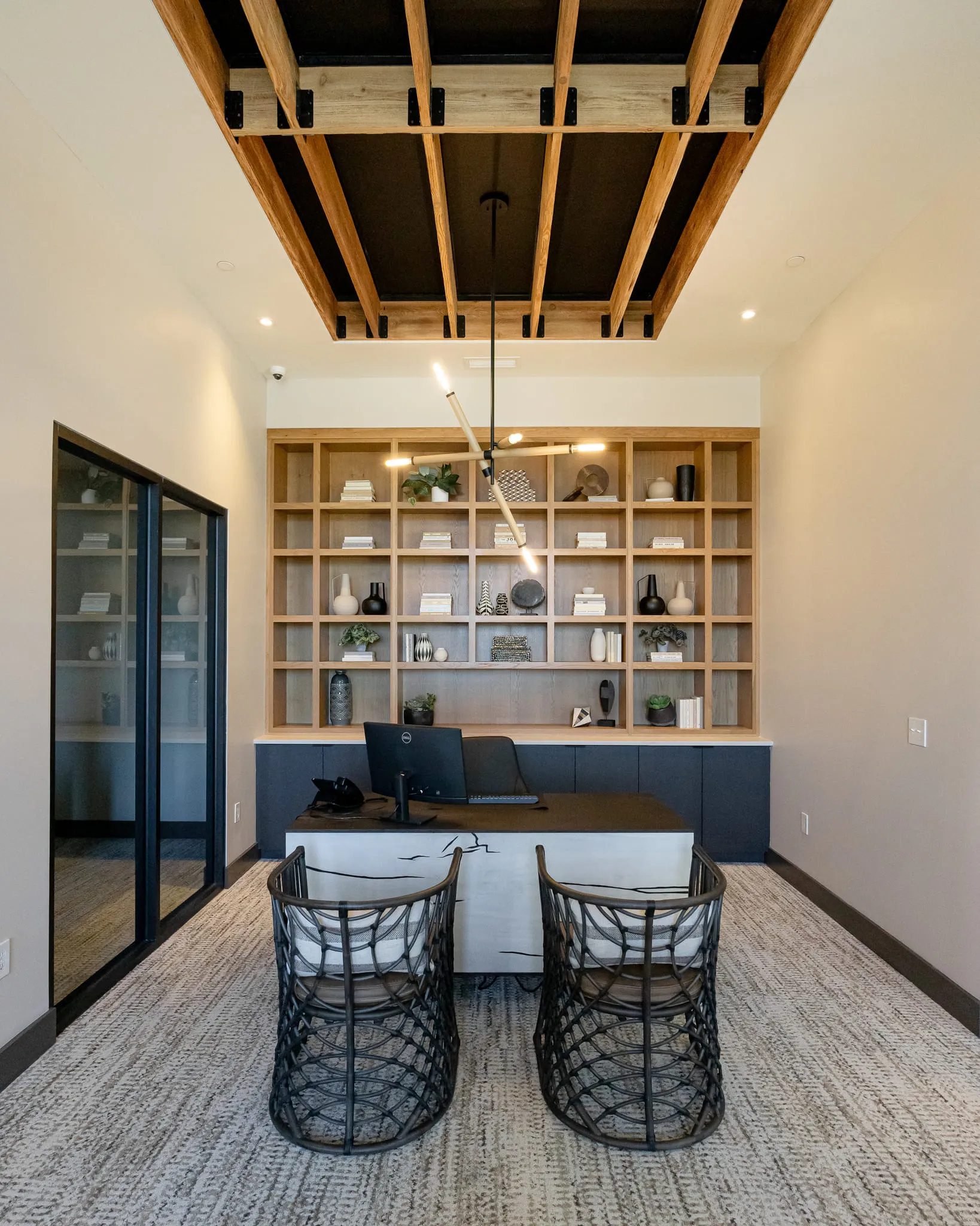 Multifamily amenity private office with an oak shelving wall, a desk and woven chairs under exposed timber, a glass door