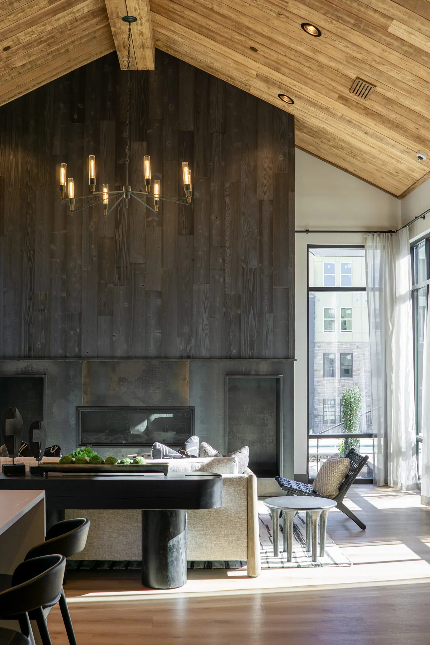 Multifamily amenity clubroom under a vaulted wood ceiling, a dark weathered plank fireplace wall and linear brass chandelier