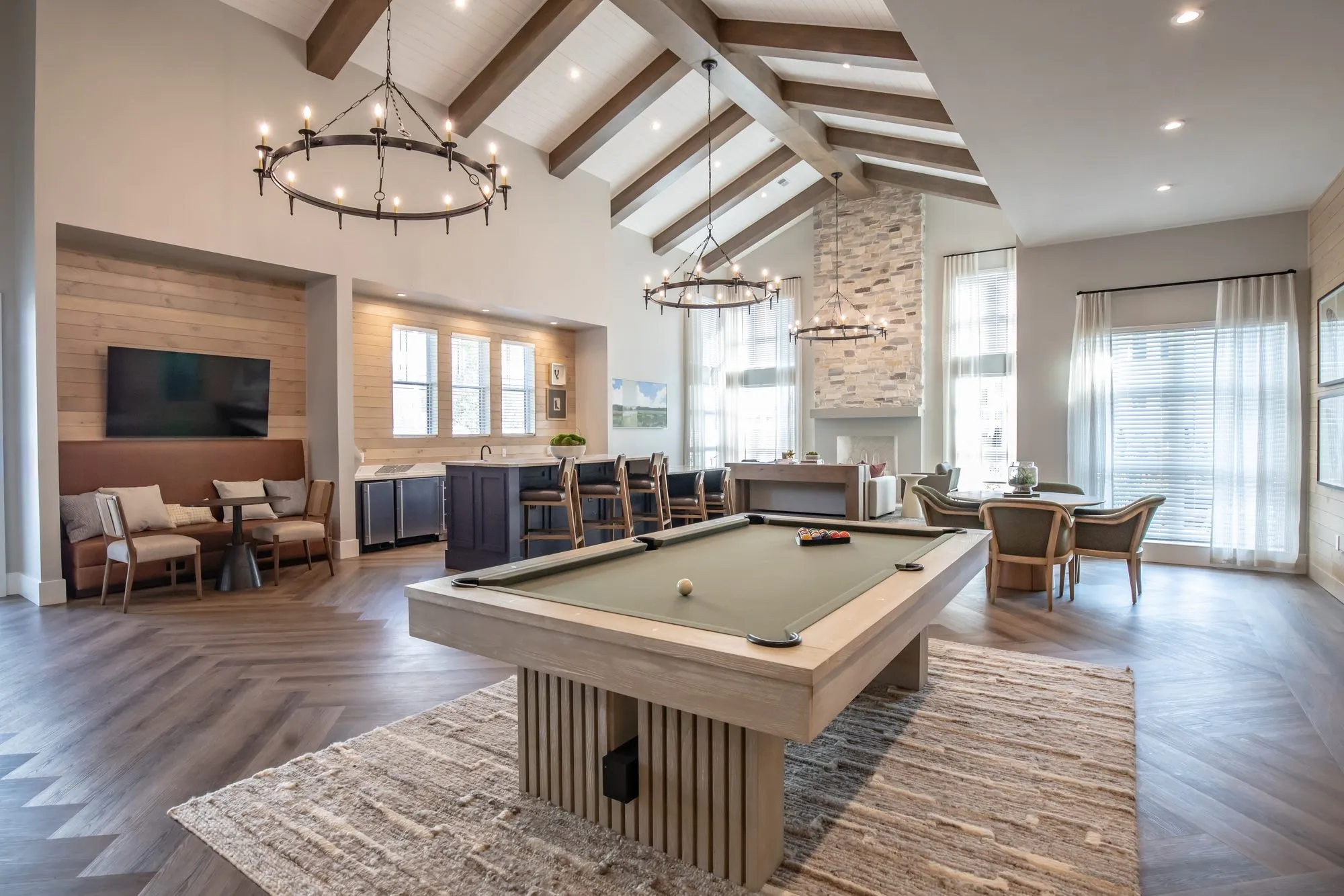 Multifamily amenity clubroom under a vaulted ceiling, pool table on a woven rug with booth seating and the bar behind
