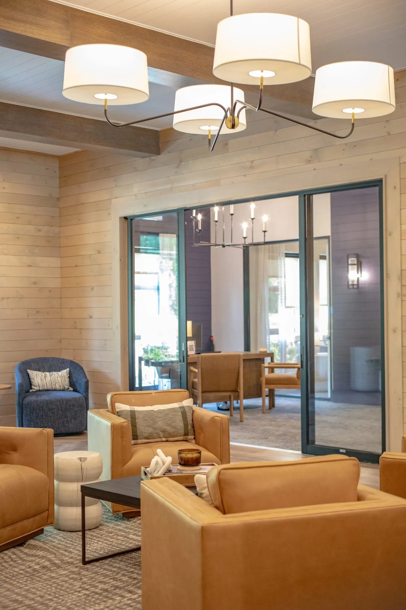 Multifamily amenity lounge with tan leather armchairs and a blue swivel chair under a drum shade chandelier, an office beyond