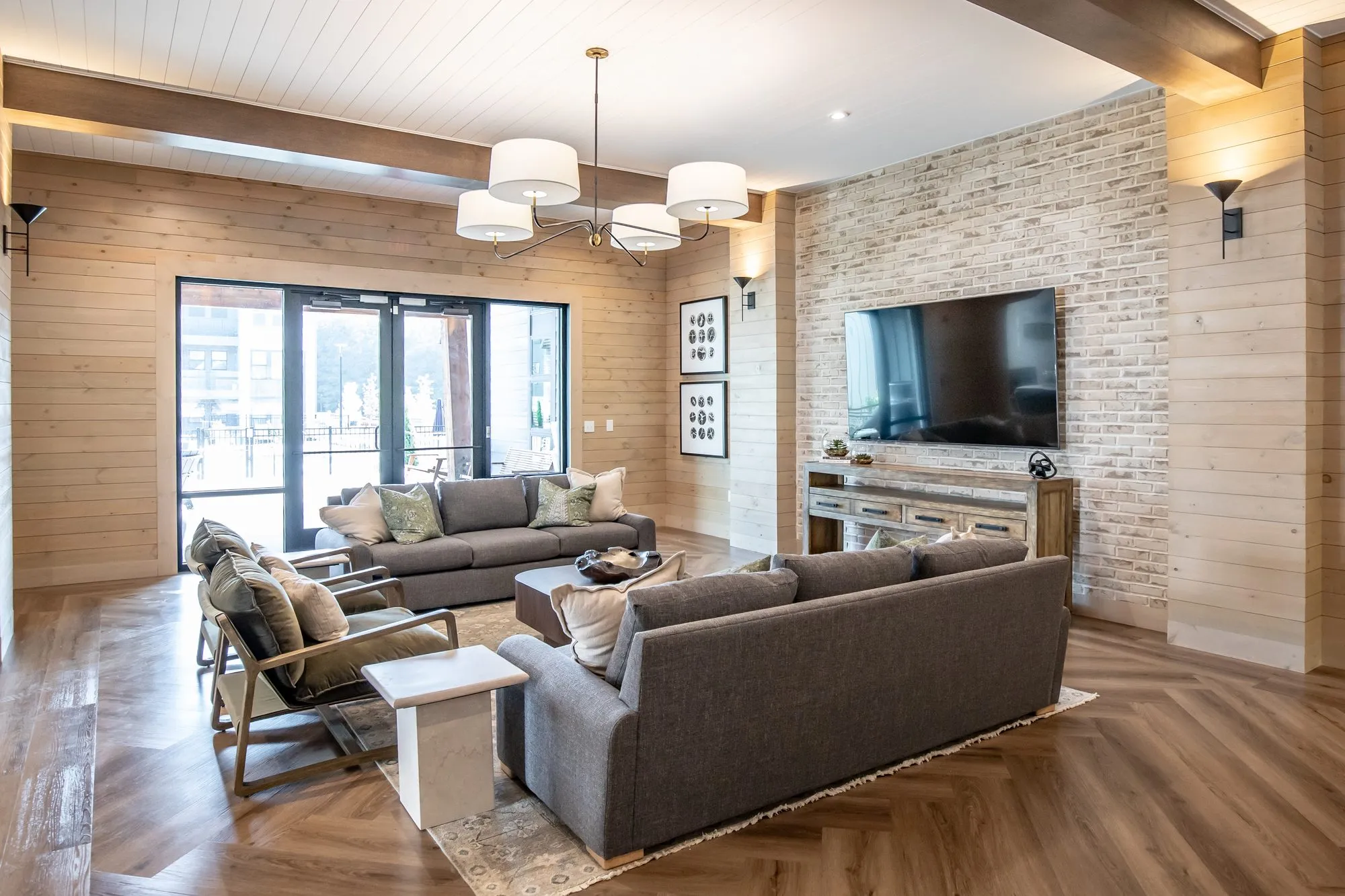 Multifamily amenity lounge with grey sofas facing a TV on a whitewashed brick wall beside glass doors