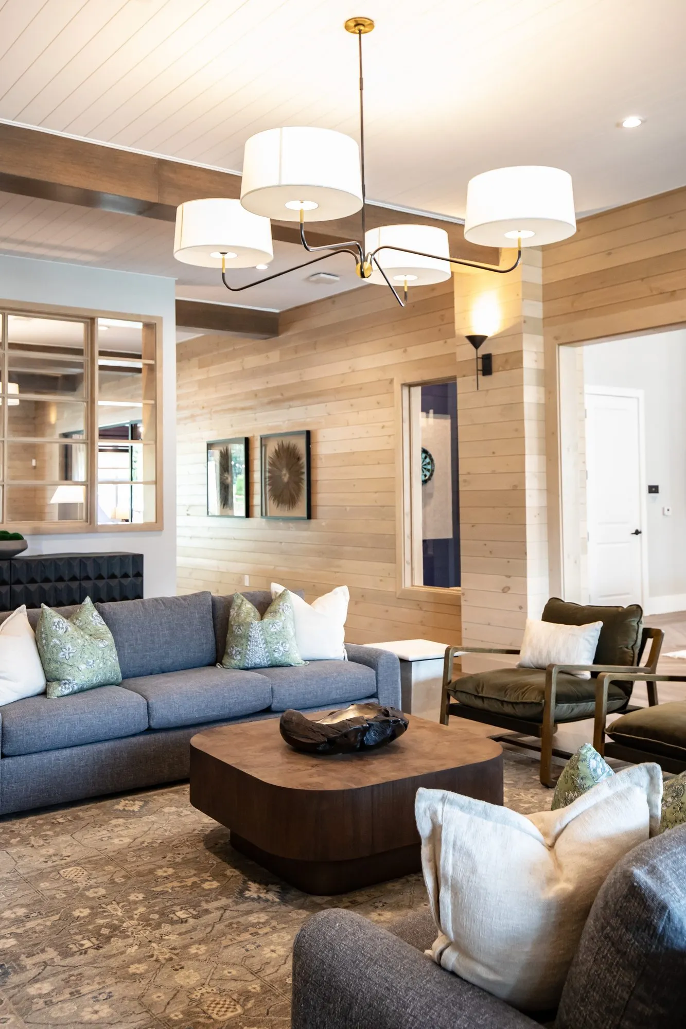 Multifamily amenity lounge looking toward the game room, grey sofa and leather armchair beneath a four shade chandelier