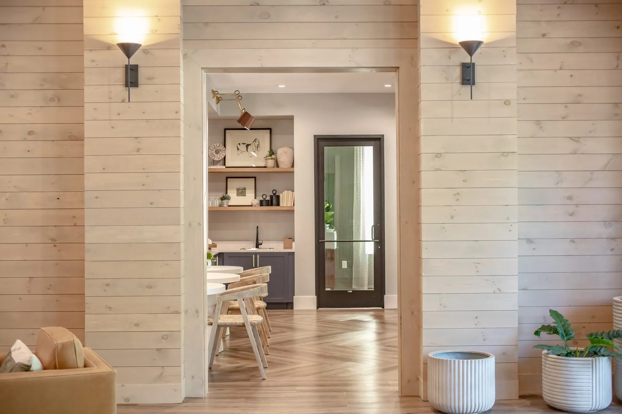 Multifamily amenity coffee bar with open shelves seen through a doorway in whitewashed plank walls between black sconces