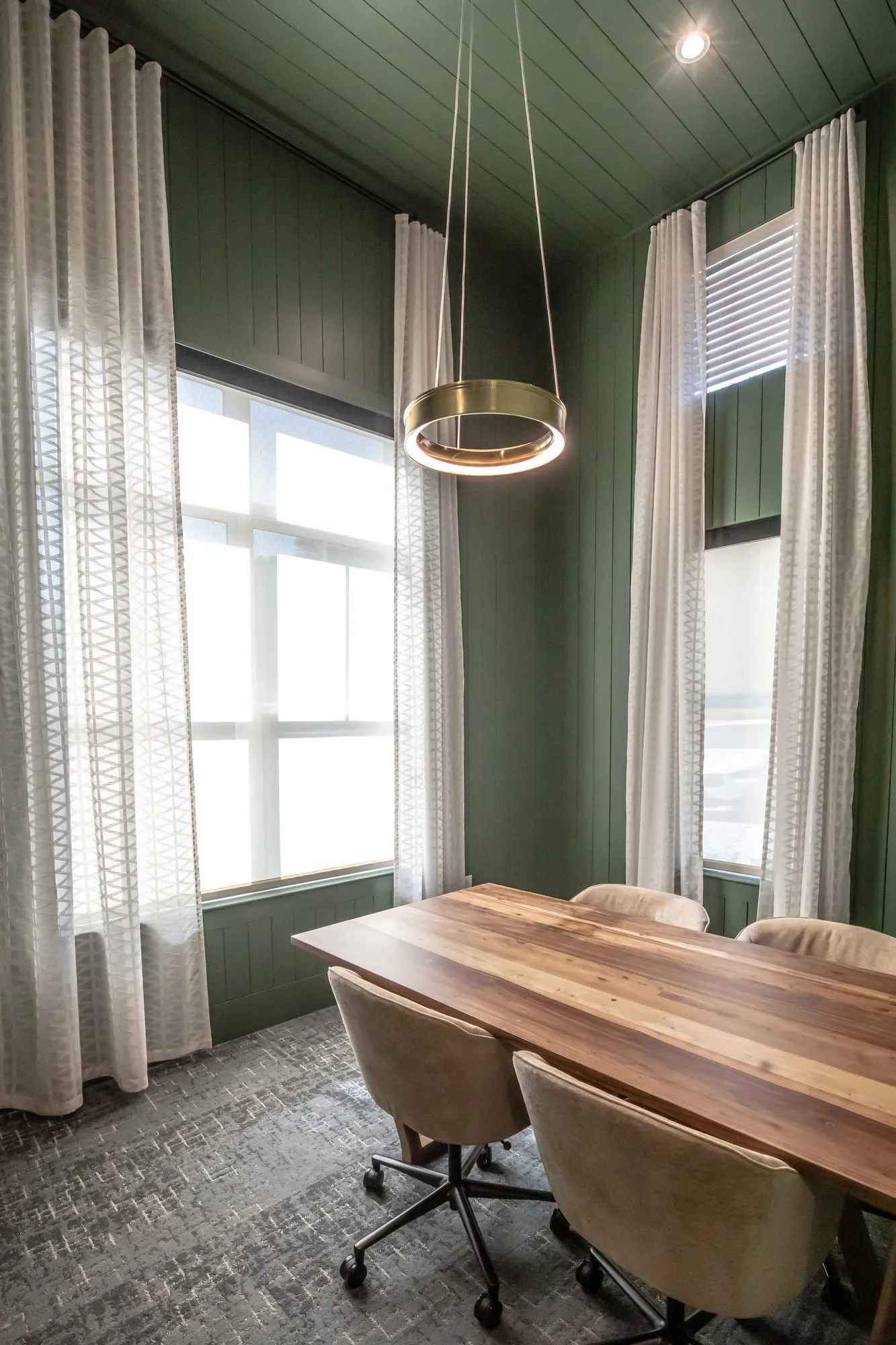 Multifamily amenity meeting room with green paneled walls, a wood table, beige chairs and a brass ring pendant by windows