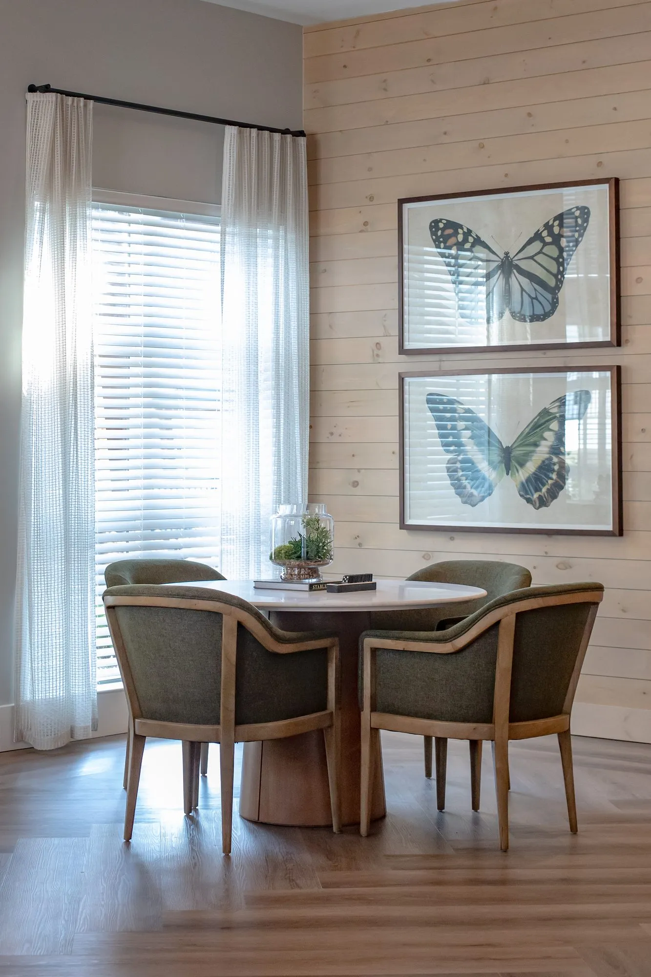 Multifamily dining corner with a round pedestal table, olive upholstered chairs and two butterfly prints on plank walls