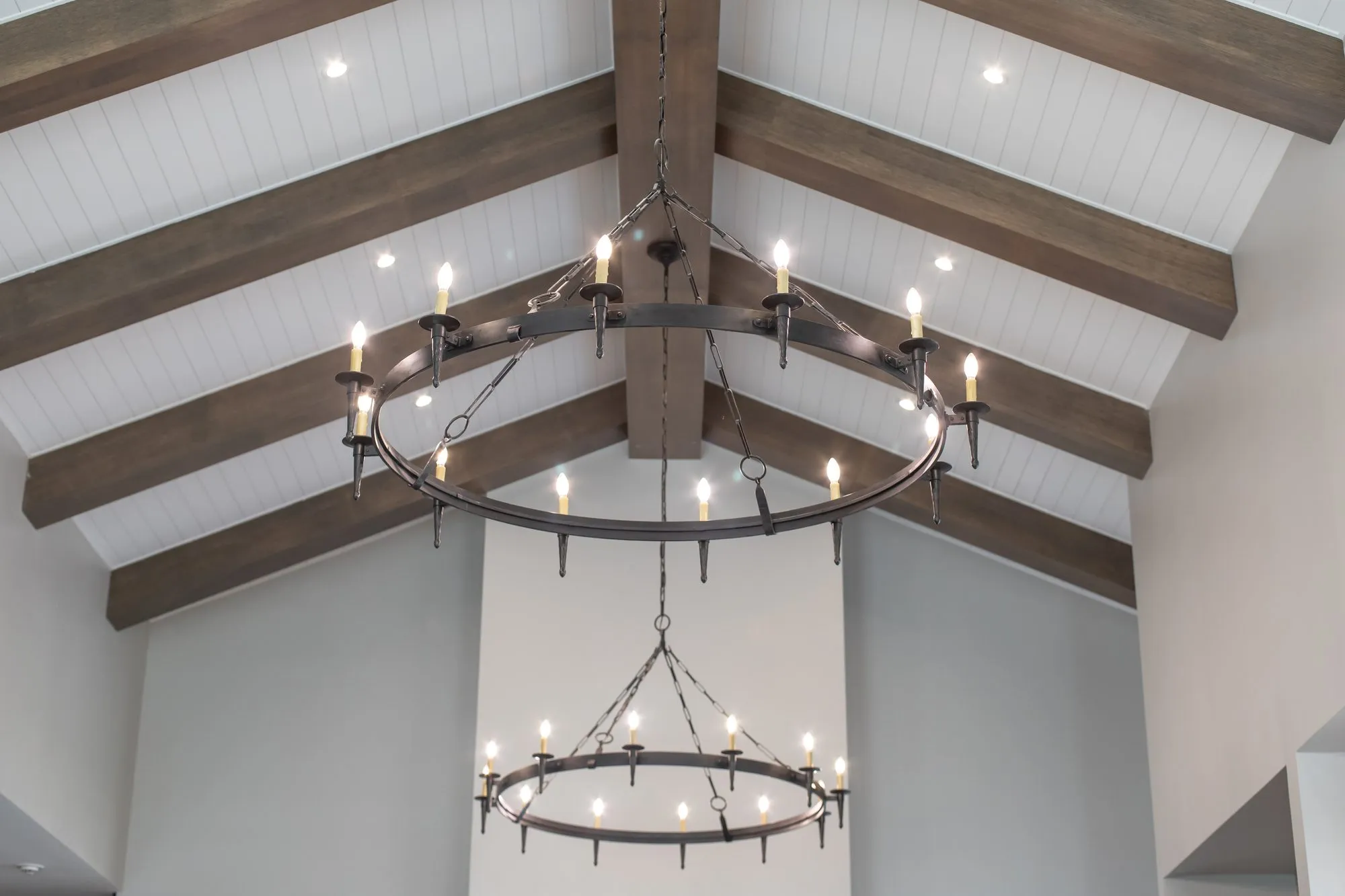 Multifamily interior detail: two tiers of black iron ring candle chandeliers from stained beams in a white vaulted ceiling