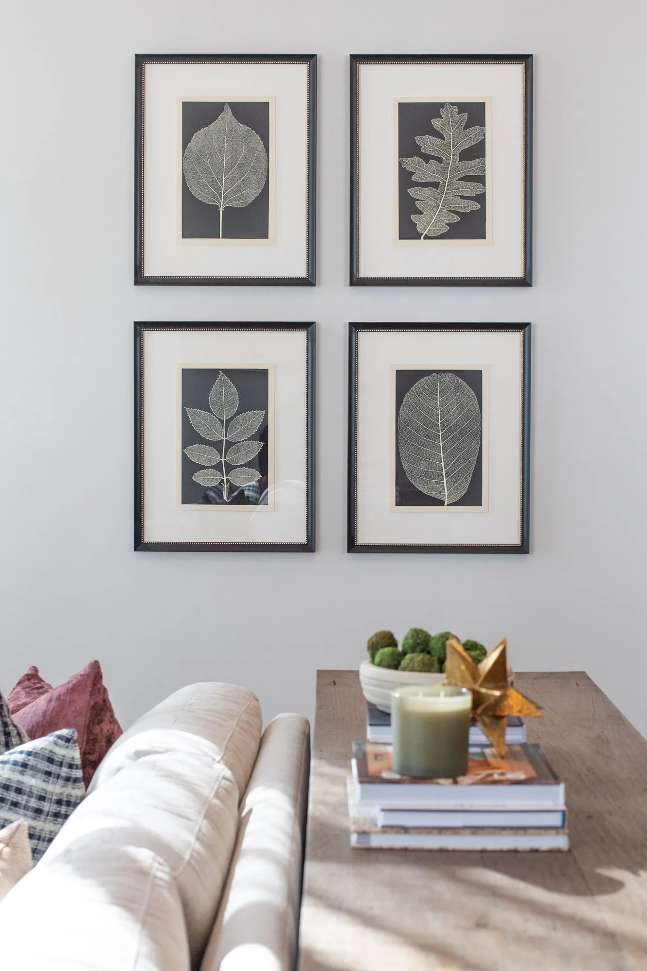 Multifamily interior detail: grid of four framed leaf prints above a cream sofa and a wood console with books and a candle