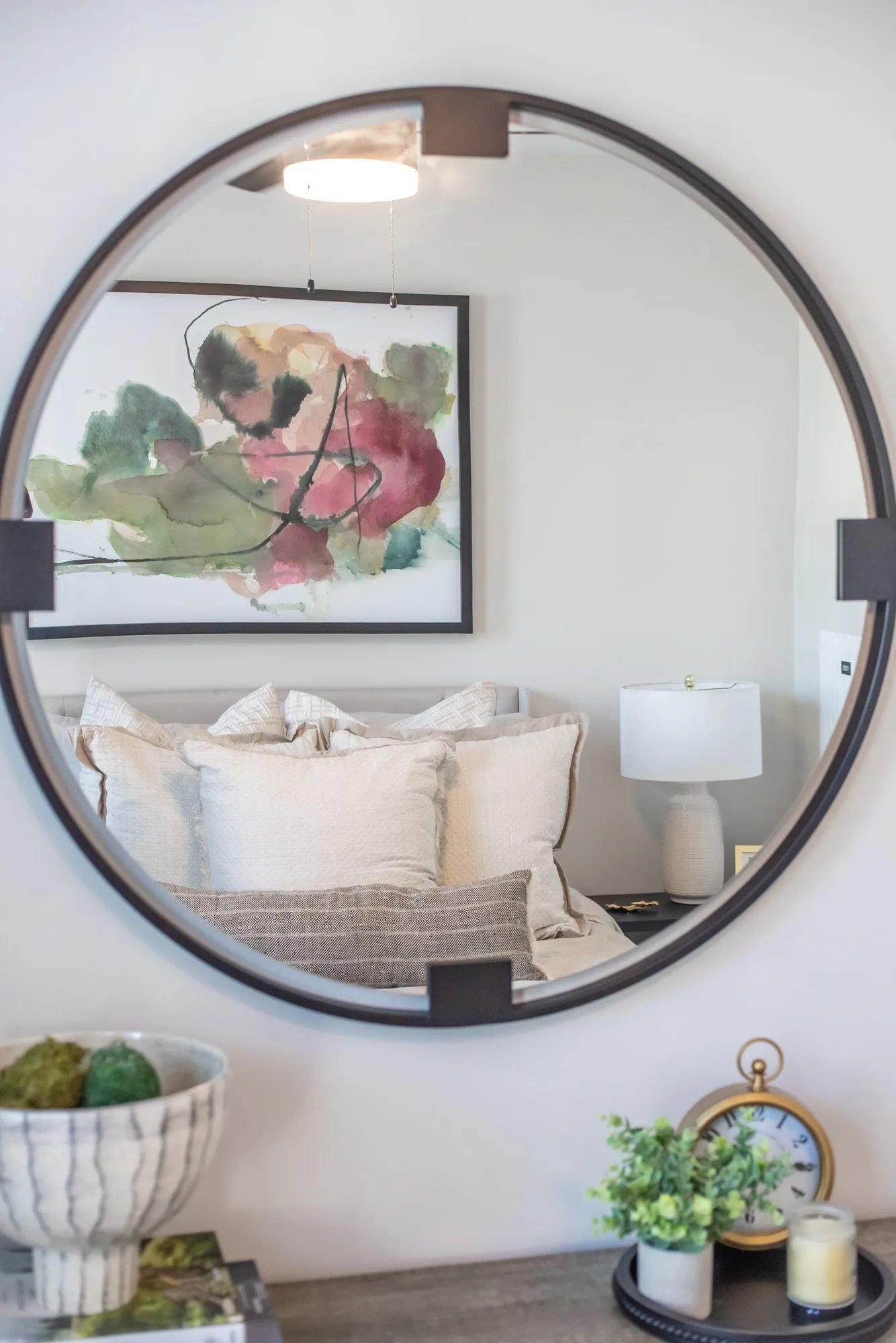 Multifamily apartment bedroom detail: round mirror reflecting the bed and a floral print, above a dresser with plants