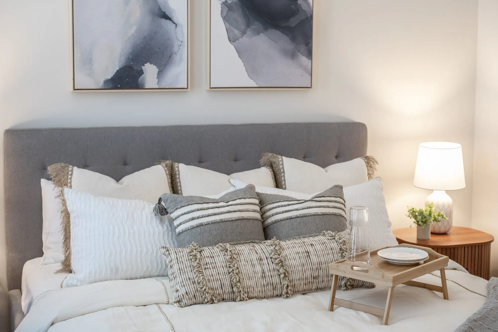 Multifamily model apartment bedroom with a grey tufted headboard, striped and fringed pillows, a bed tray and abstract prints
