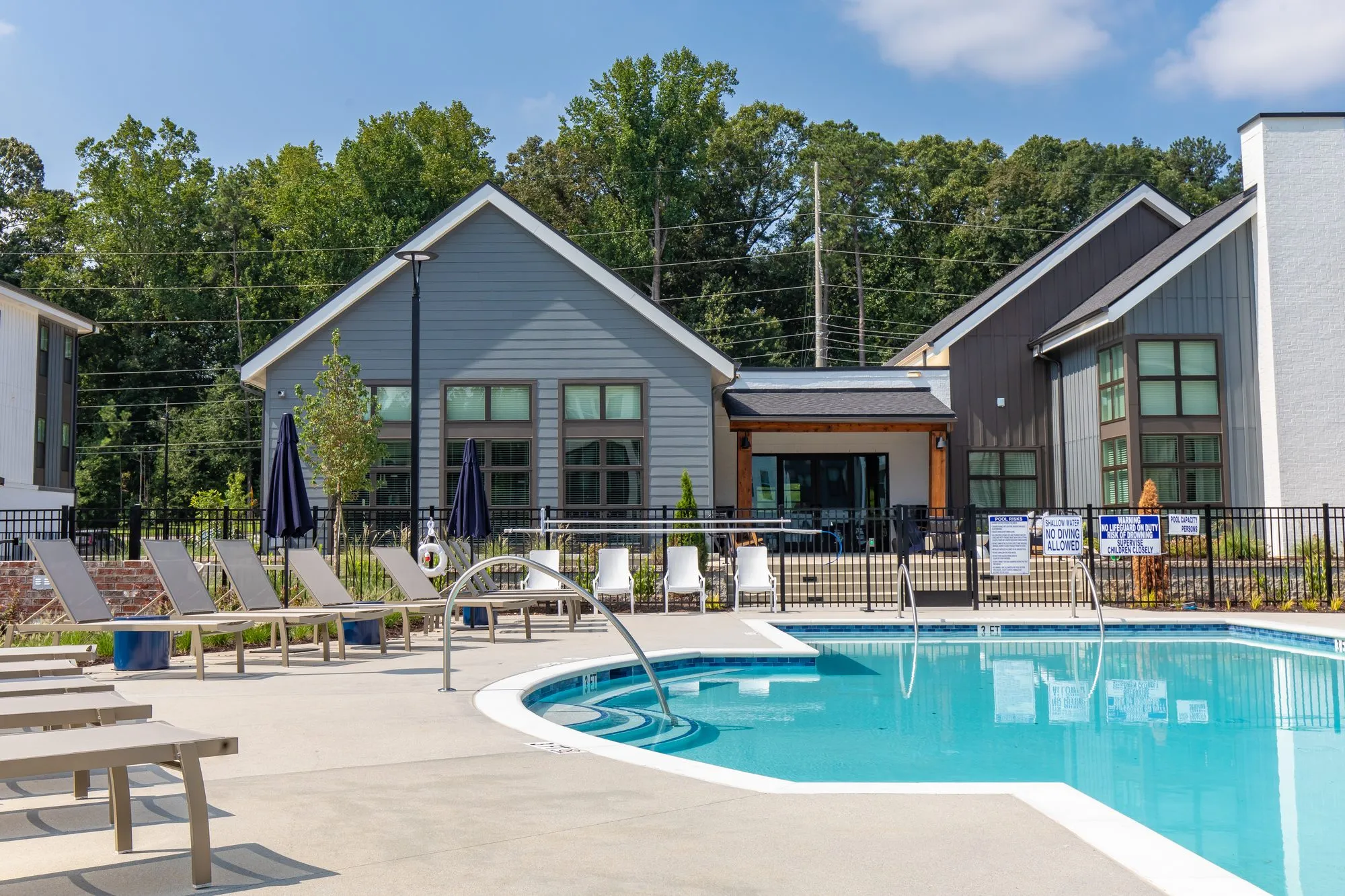 Multifamily amenity pool deck with grey loungers and blue umbrellas beside the clubhouse with grey and white gabled siding