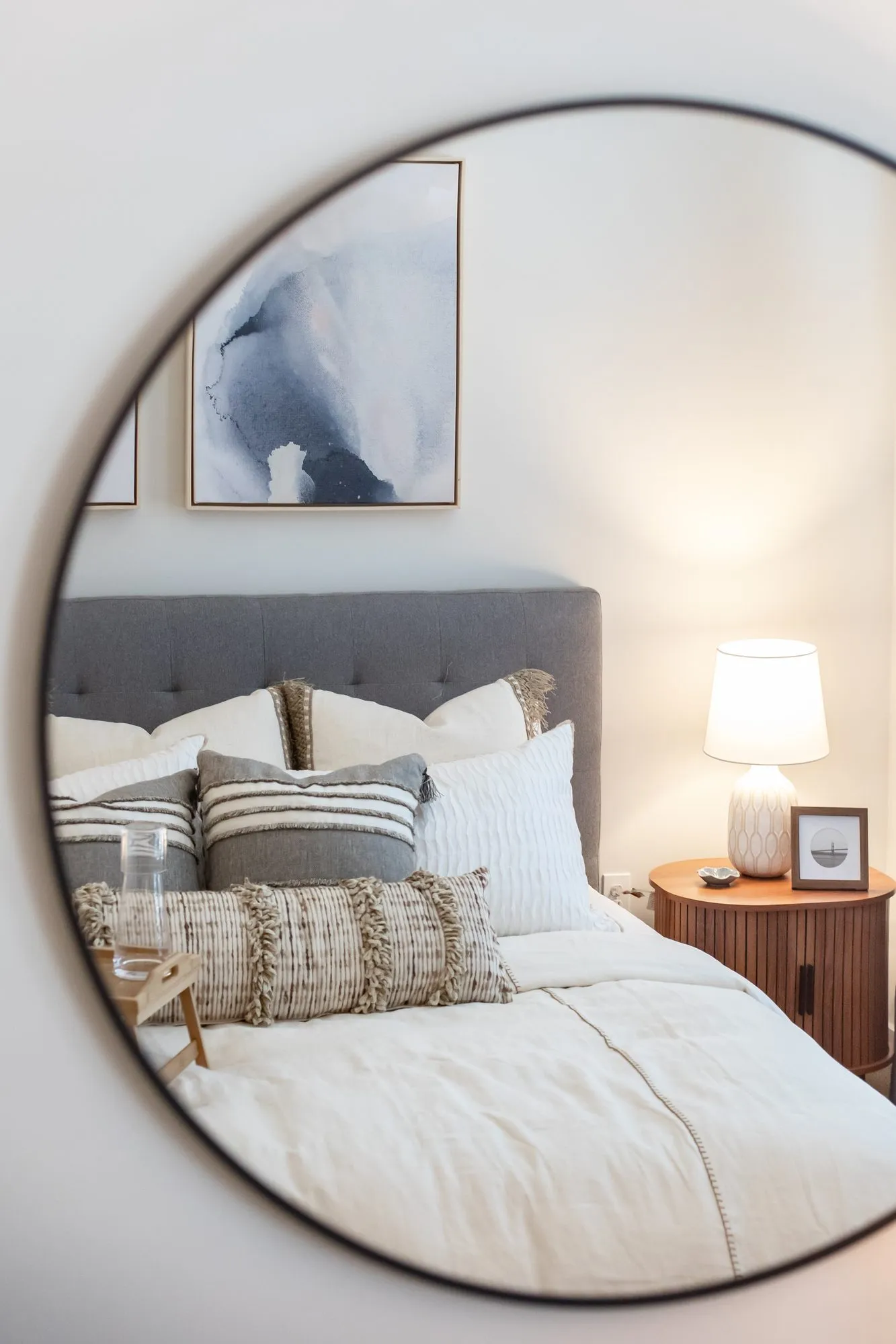 Multifamily apartment bedroom detail: round mirror reflecting a bed with a grey tufted headboard and a ribbed wood nightstand