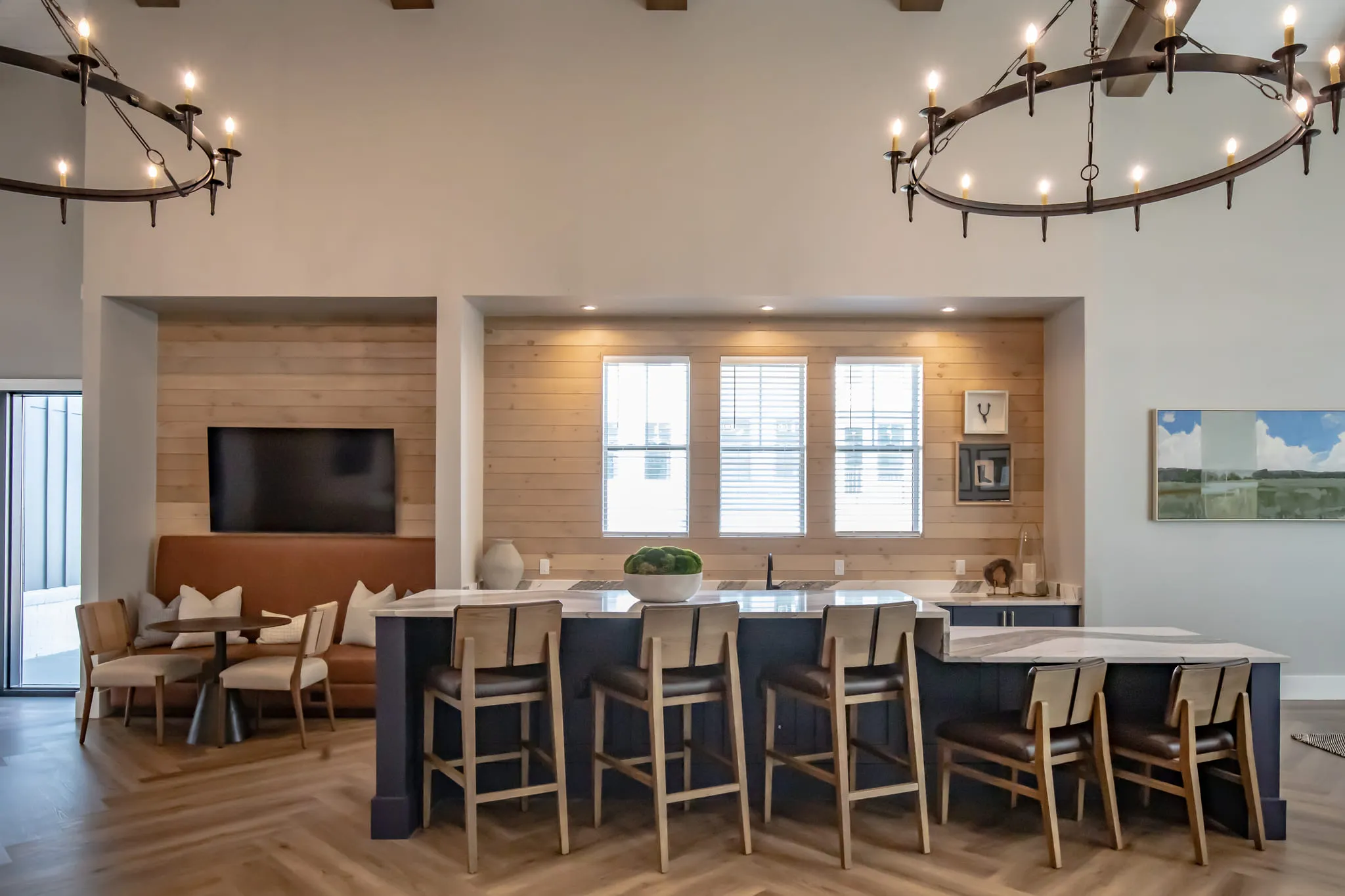Multifamily amenity clubroom bar with a navy counter and wood stools below a ring chandelier and a wall-mounted screen