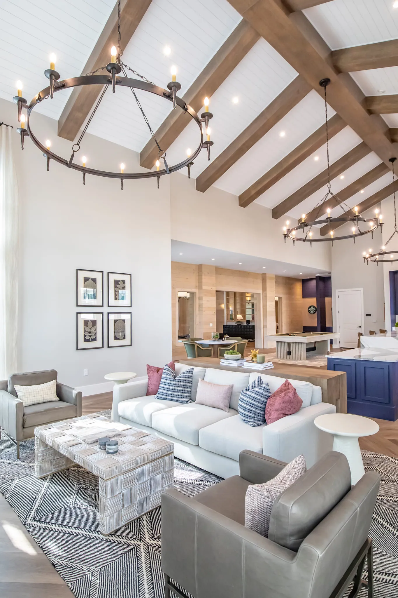 Multifamily amenity clubroom with white sofas and an iron ring chandelier below the beamed ceiling, the kitchen island beyond