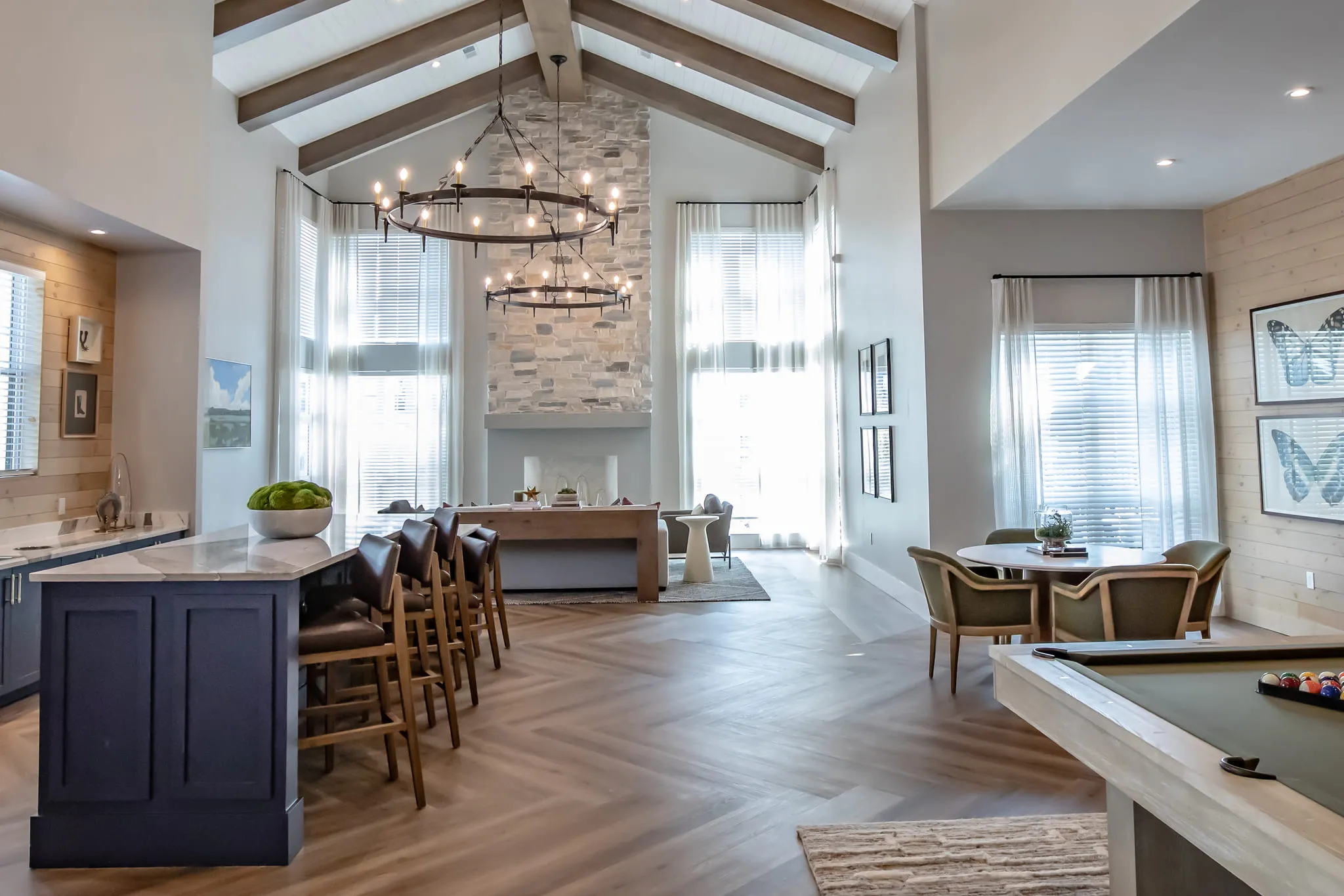 Multifamily amenity clubroom with a navy island and leather barstools under iron ring chandeliers, a stone chimney beyond