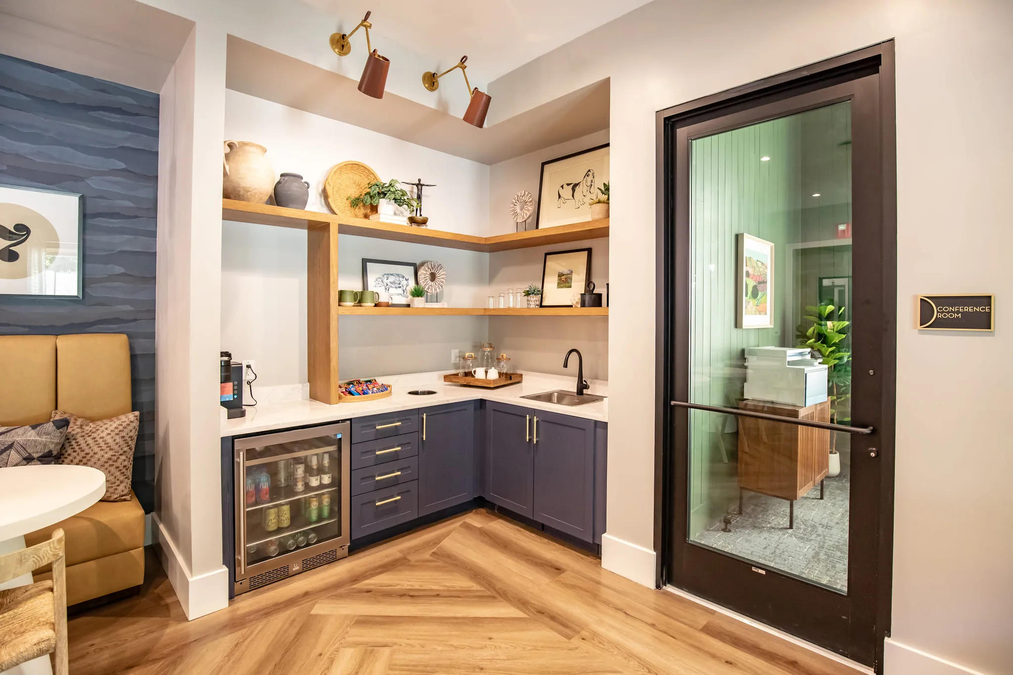 Multifamily amenity coffee bar with navy cabinetry and open oak shelving on herringbone floors beside a glazed door