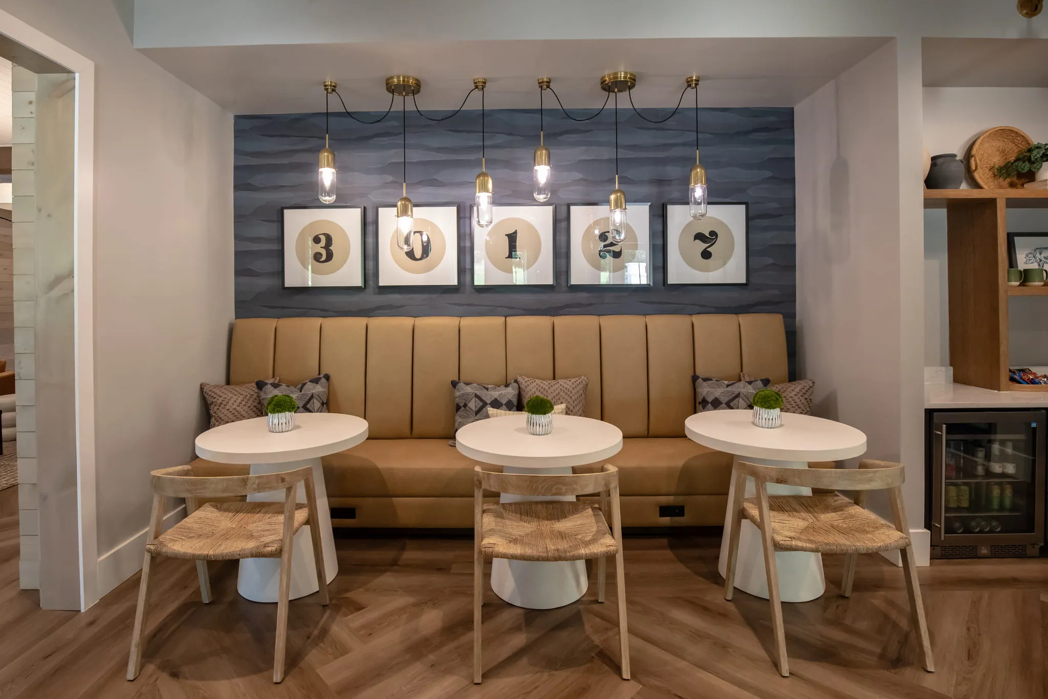 Multifamily amenity coffee nook with a tan channel-tufted banquette and round tables below brass pendants and numbered art