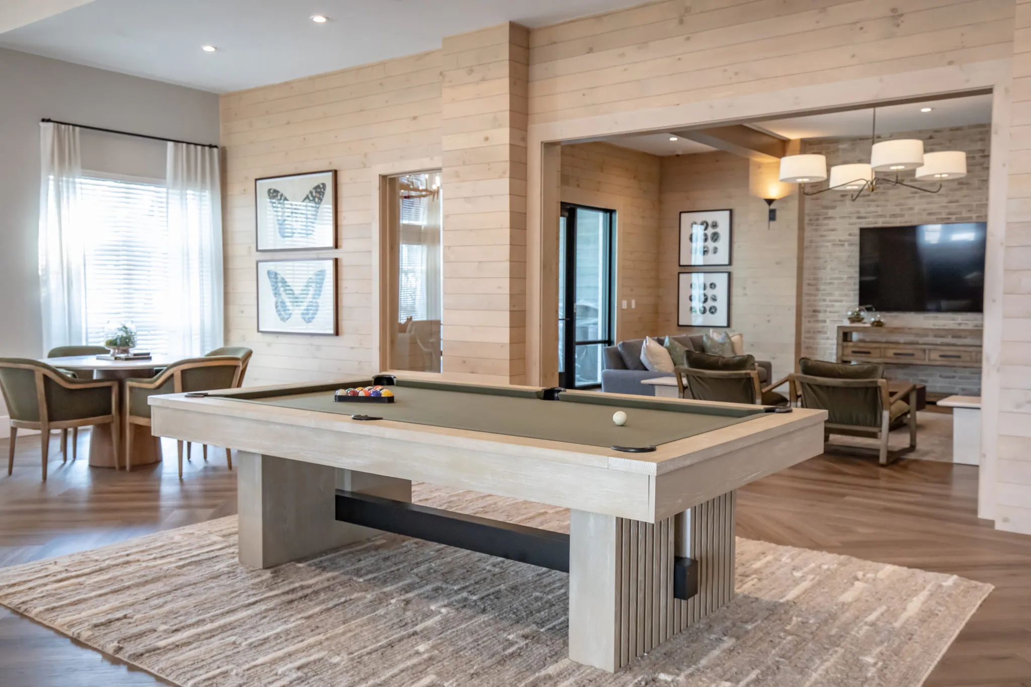Multifamily amenity game room with a pool table on a patterned rug between framed butterfly prints, the lounge beyond