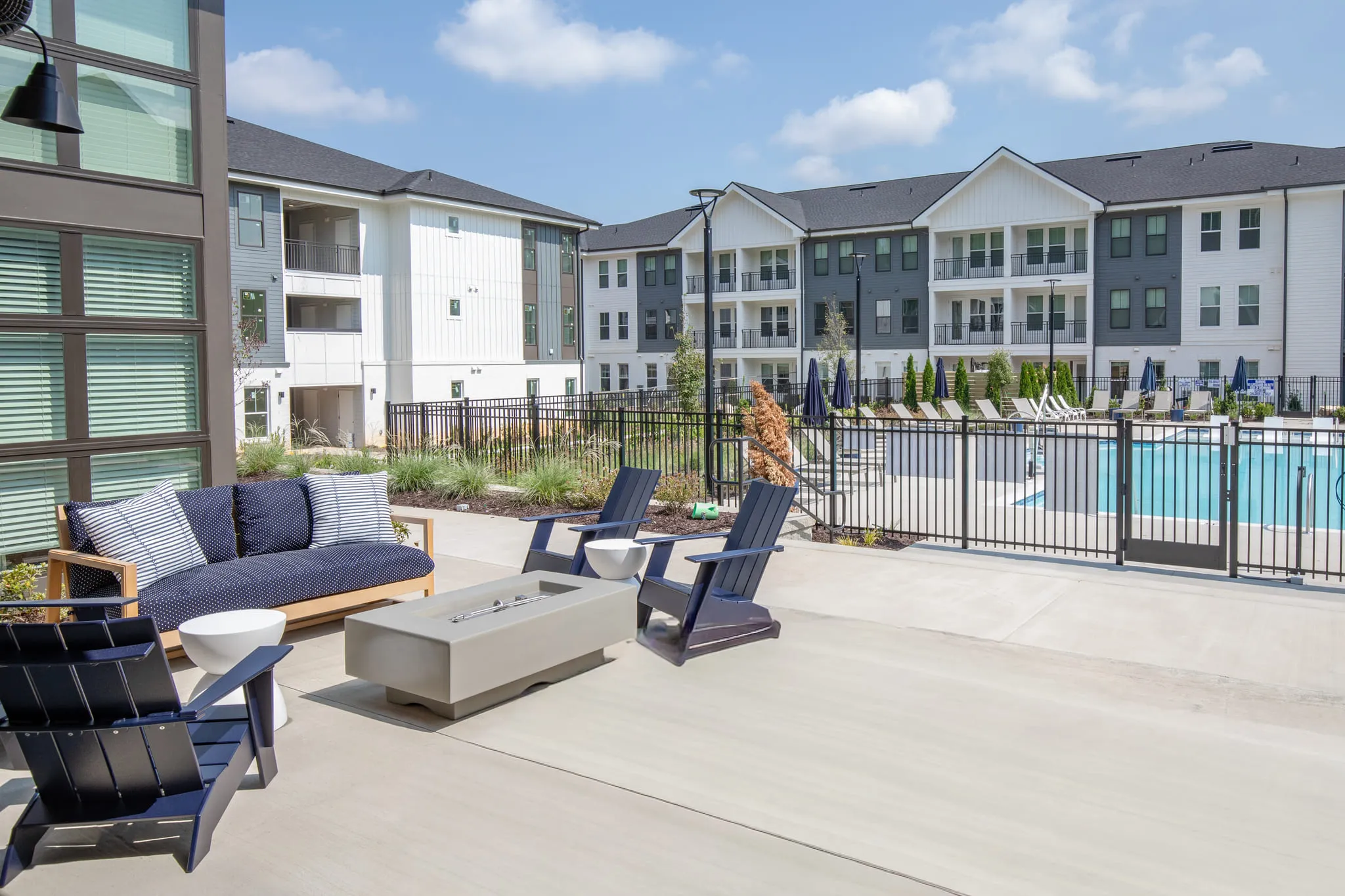 Multifamily amenity terrace with a navy outdoor sofa and Adirondack chairs around a concrete fire table, the pool beyond