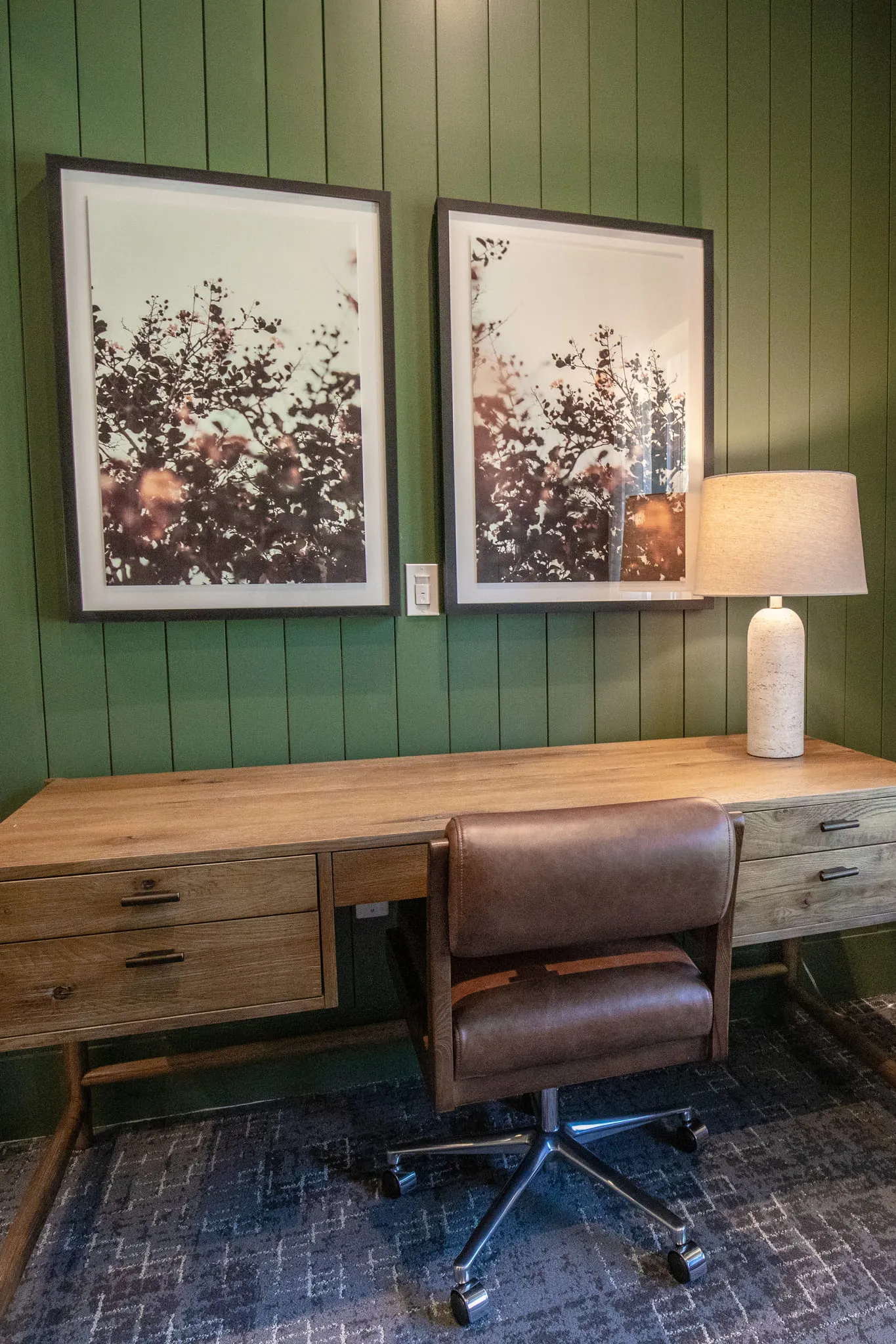 Multifamily amenity work nook with a wood desk and leather chair against green tongue-and-groove paneling below two prints