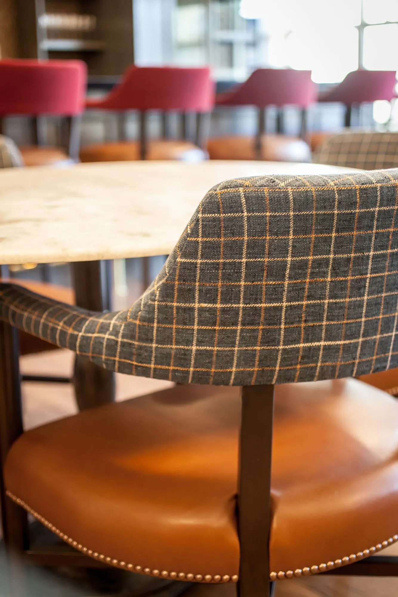 Senior living interior detail: plaid dining chair with an orange leather seat and nailheads at a marble table, red barstools