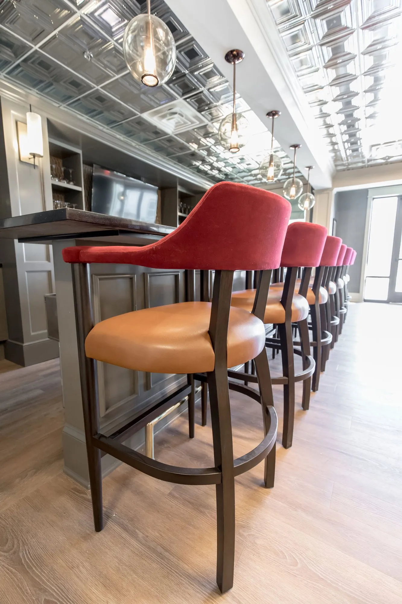 Senior living bar: low angle along barstools with red backs and cognac seats, glass pendants and pressed tin ceiling above