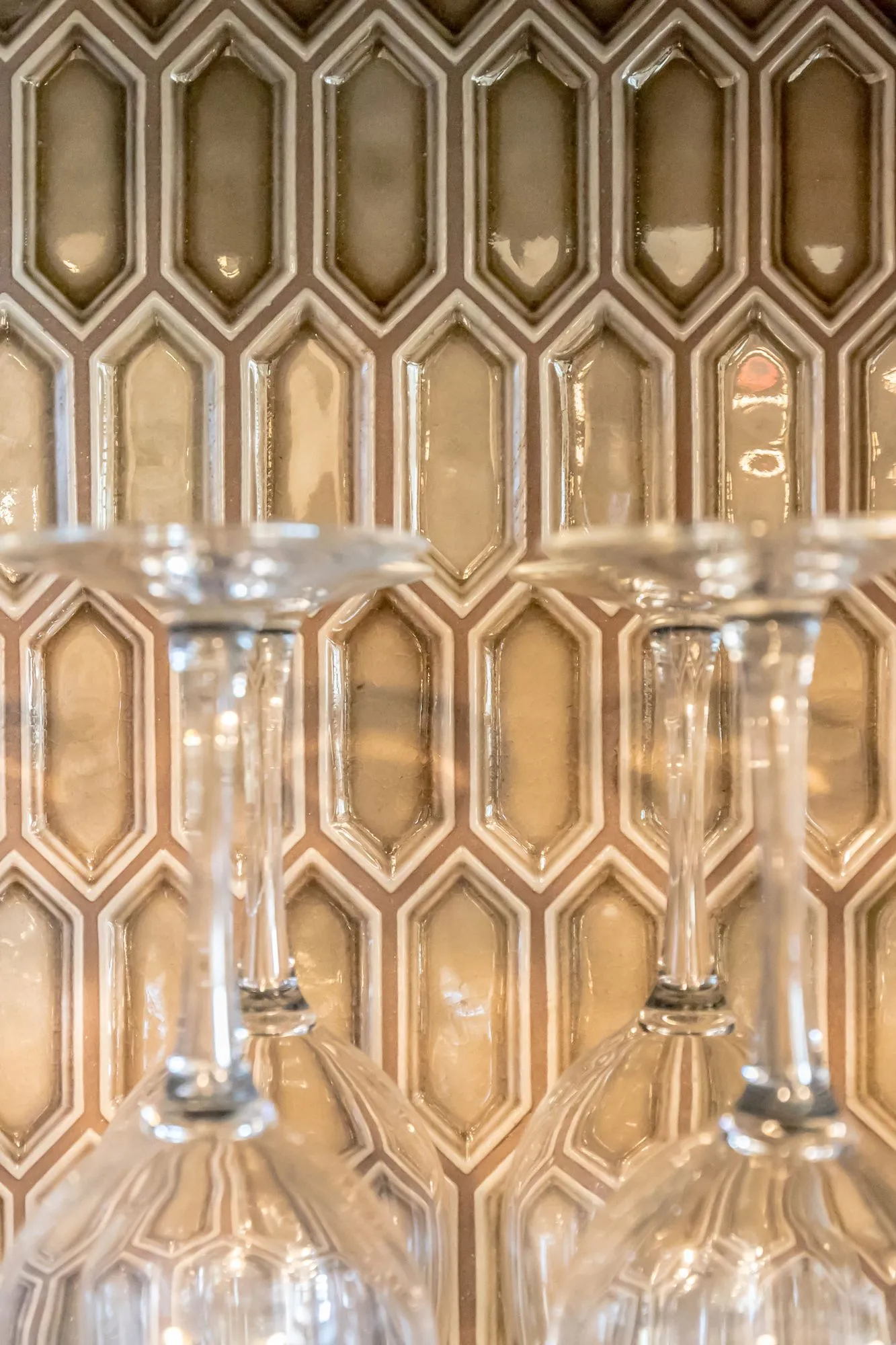 Senior living interior detail: stemmed wine glasses hanging upside down before a gold elongated hexagon tile backsplash