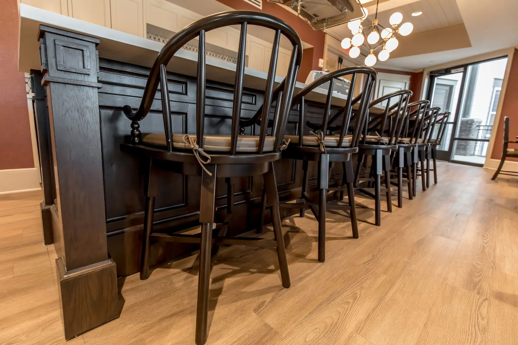 Senior living kitchen, low angle along a dark wood island lined with spindle back barstools, a globe chandelier beyond