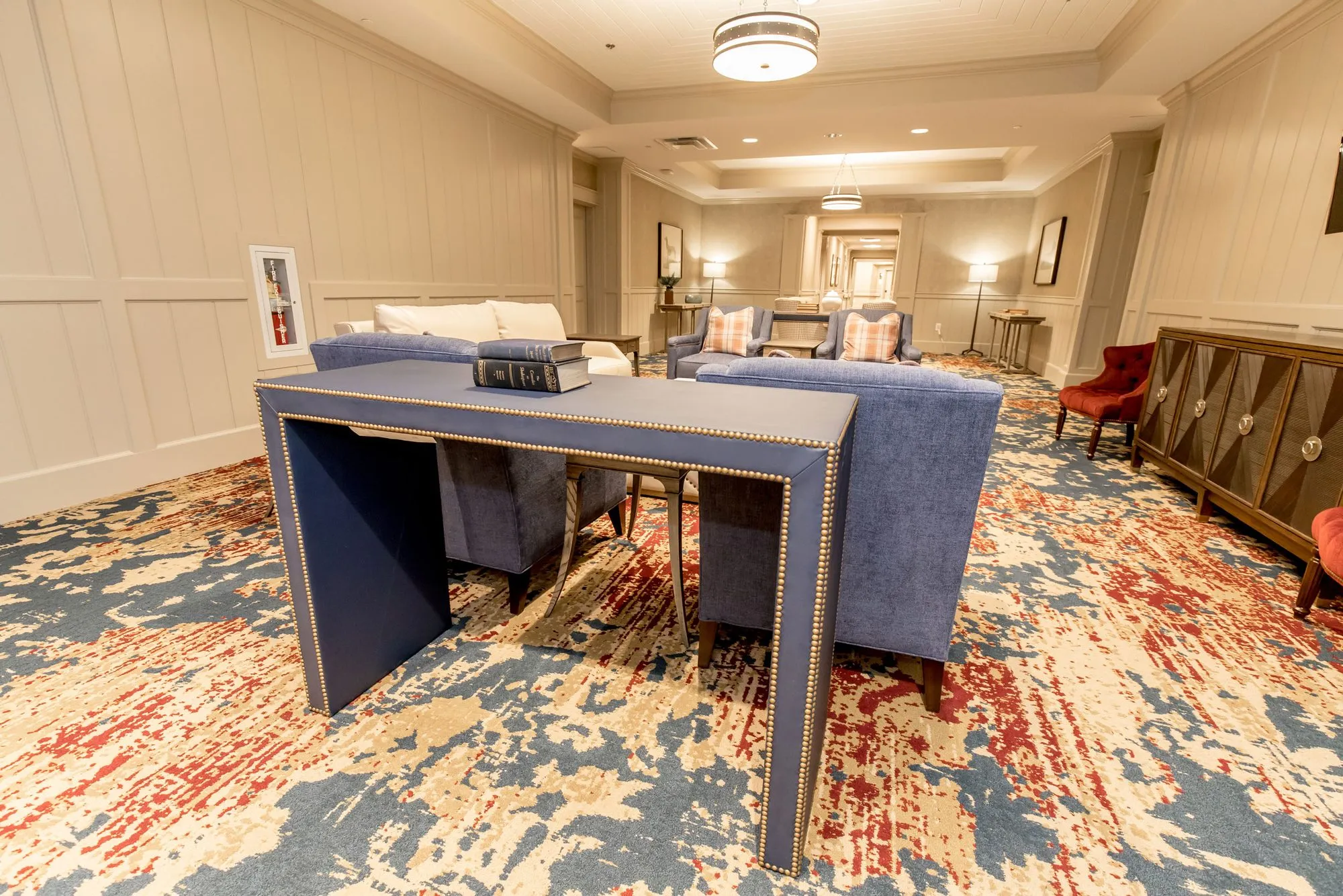 Senior living lounge with a blue, red and gold patterned carpet, a navy nailhead console table and blue sofas