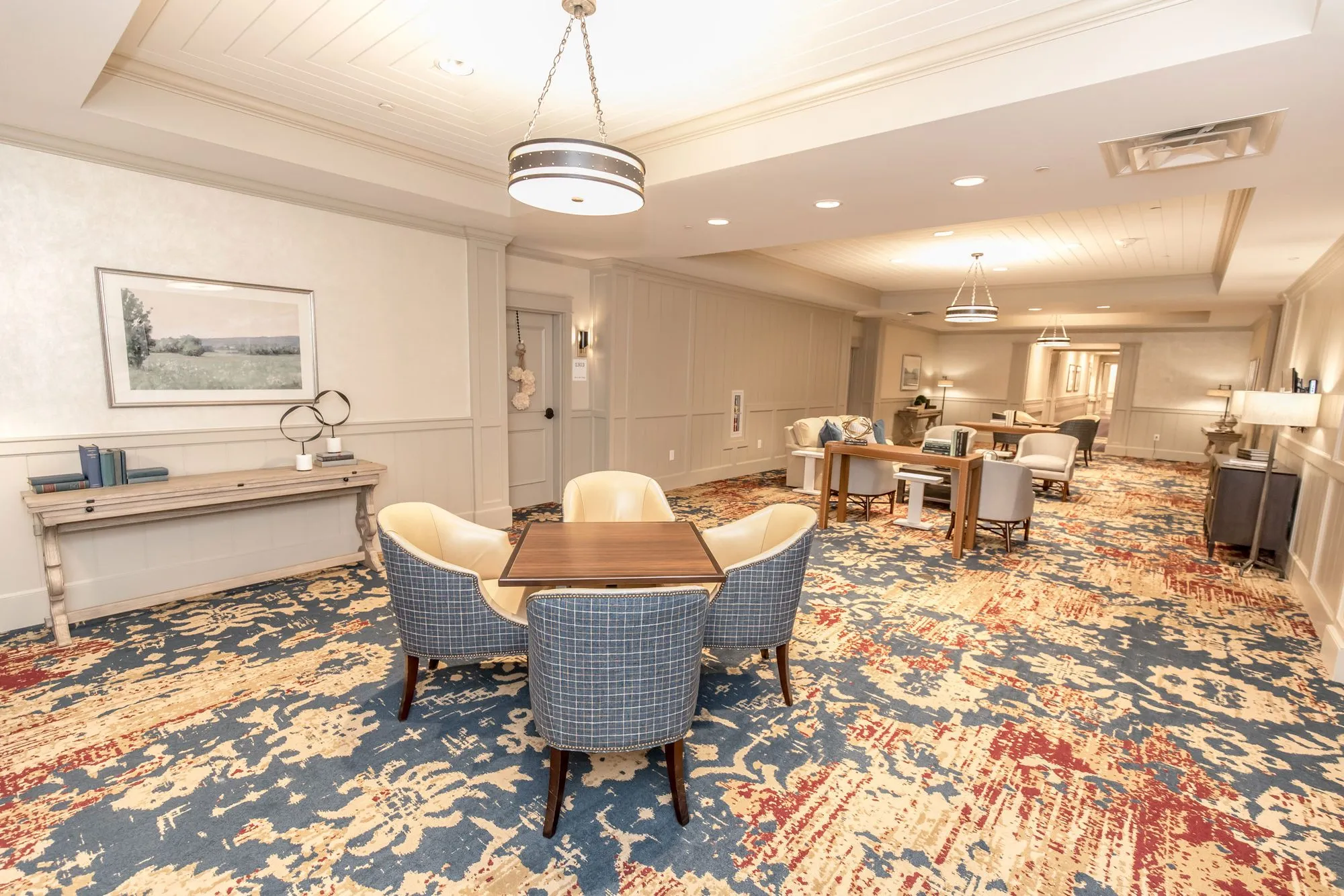 Senior living lounge with a card table and blue plaid chairs on patterned carpet, more seating under drum pendants beyond