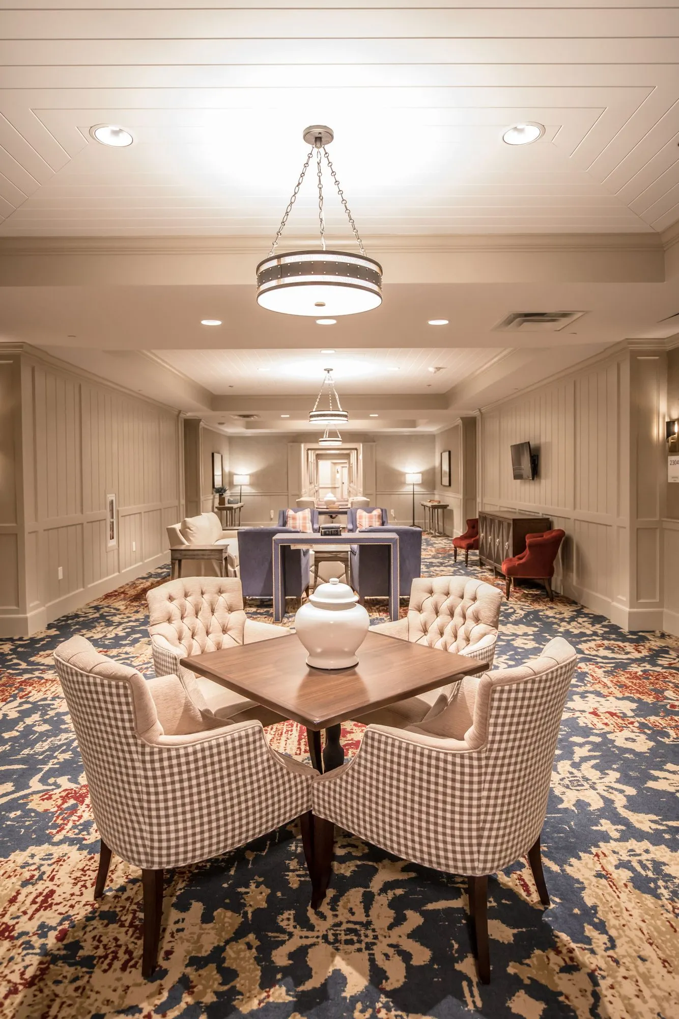 Senior living interior detail: square table with gingham tufted chairs and a white vase on a blue patterned carpet