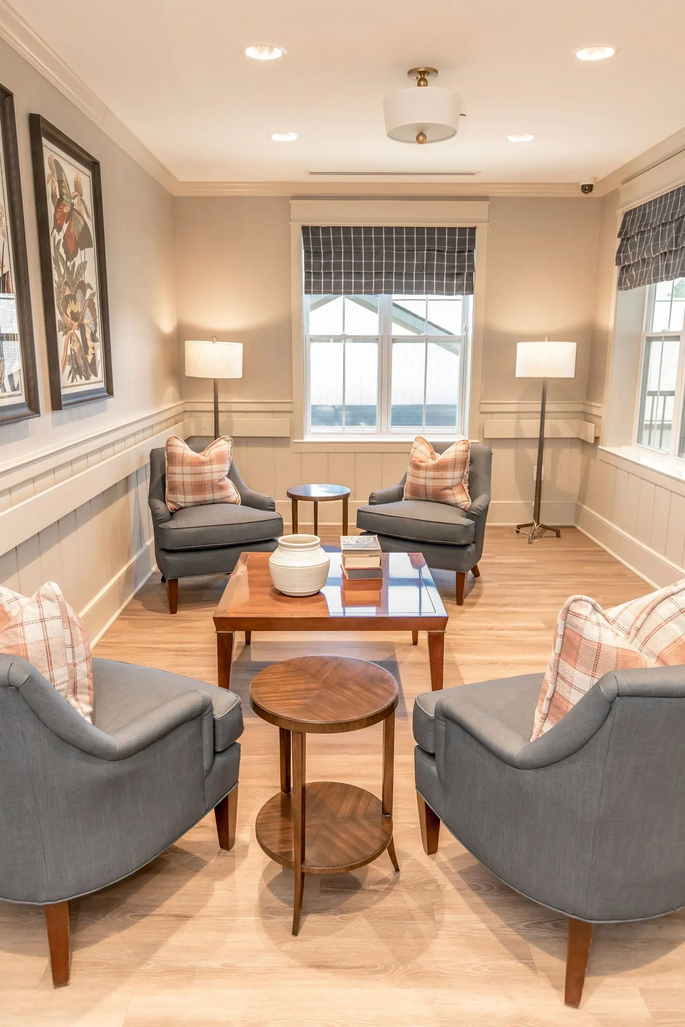 Senior living sitting room with grey armchairs and plaid pillows around a wood coffee table, botanical prints