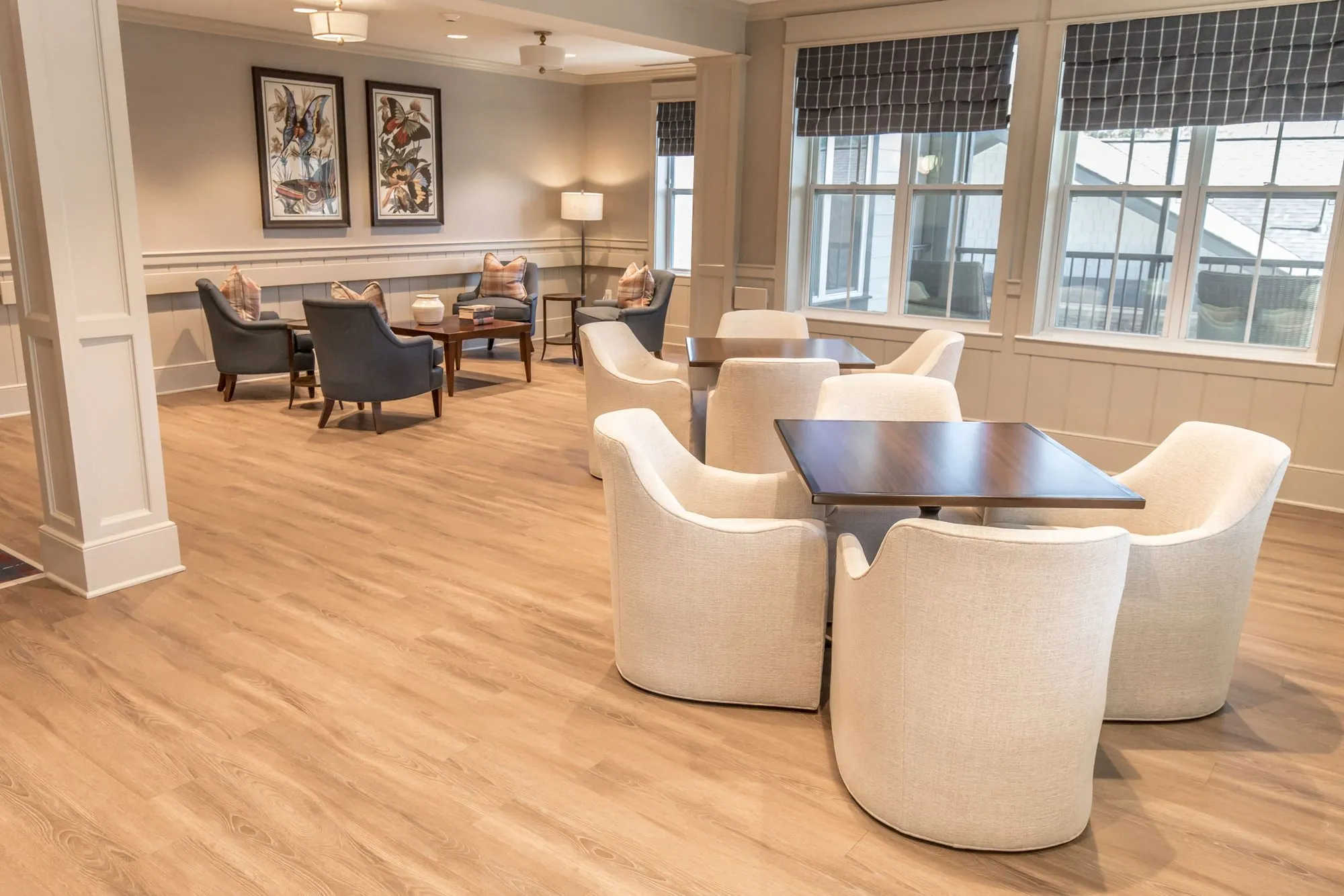 Senior living sitting area with cream barrel chairs at dark square tables by windows with plaid roman shades