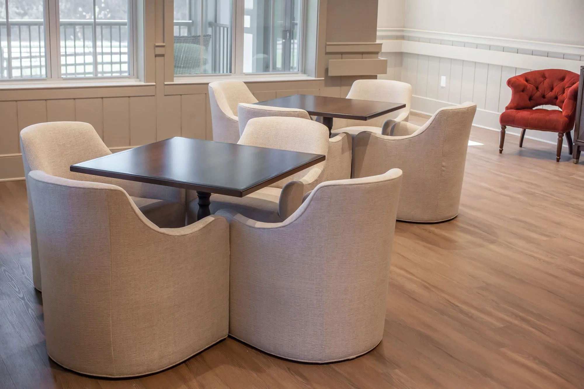Senior living interior: cream upholstered barrel chairs around dark square tables, a red tufted armchair by the wall