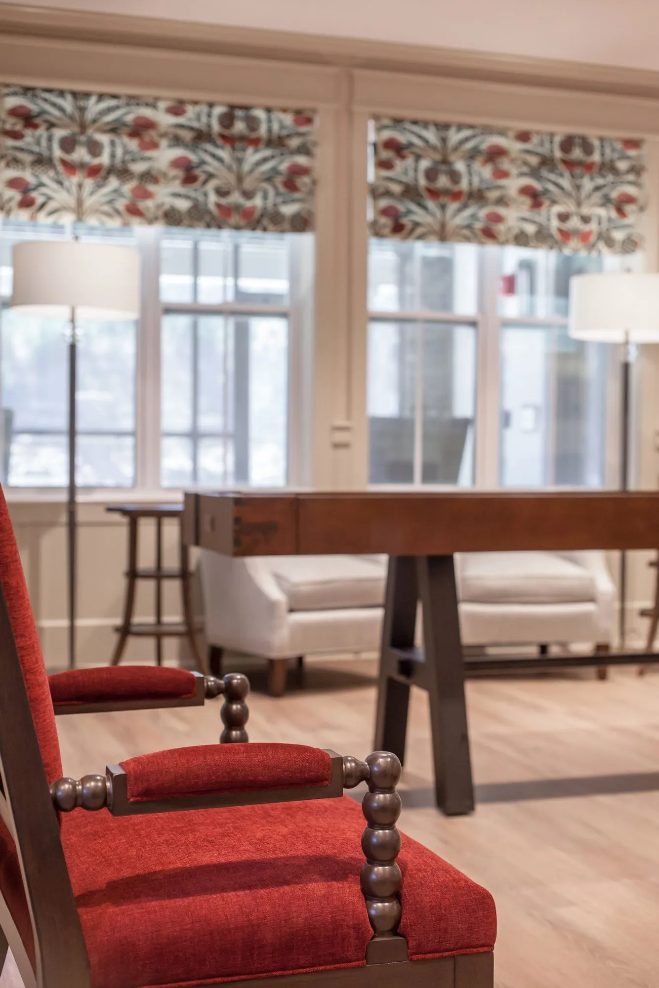 Senior living interior detail: red chair with spindle arms beside a wood table, a cream sofa and floral roman shades behind