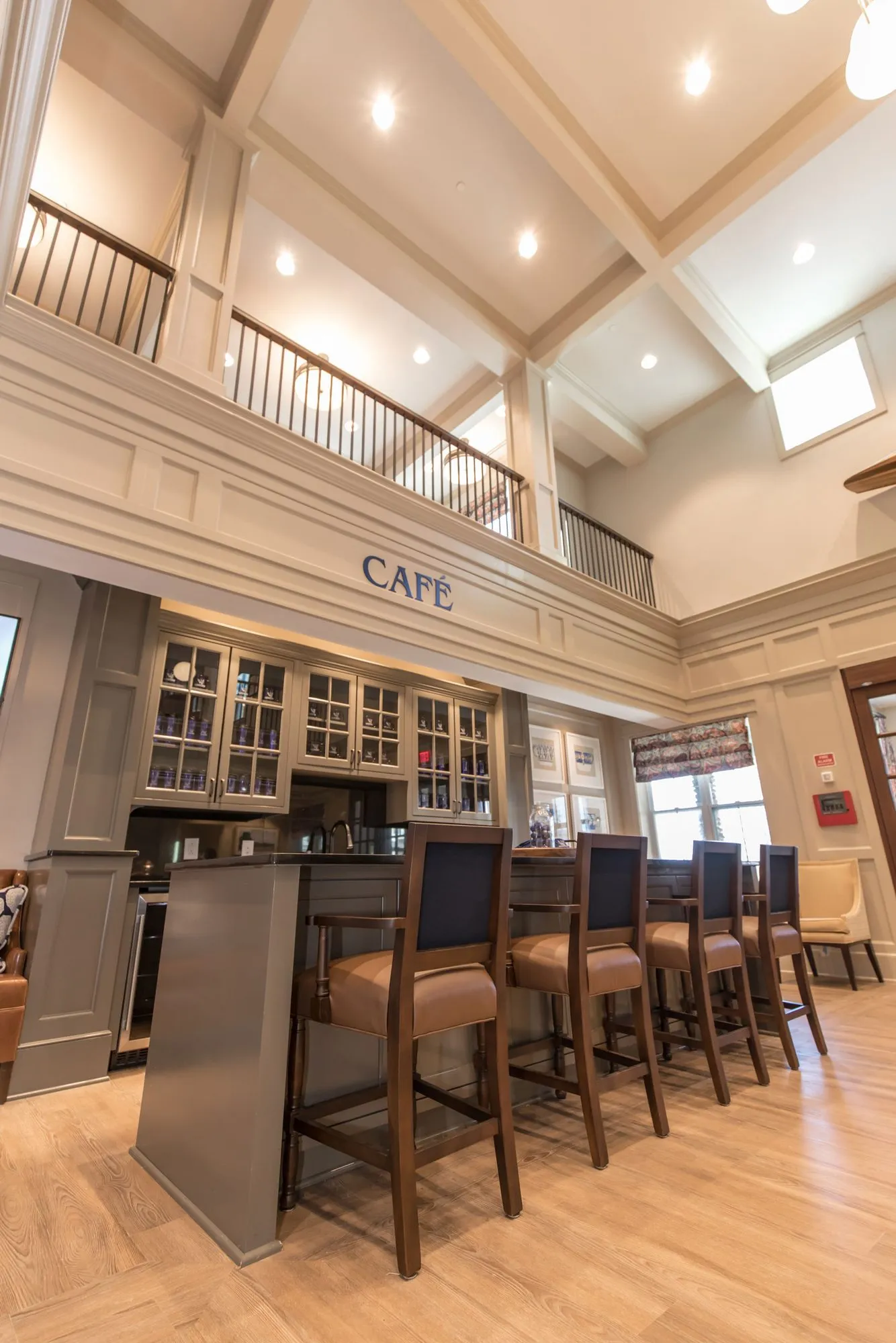 Senior living cafe under a gallery balcony, leather barstools at a counter and glass front cabinets