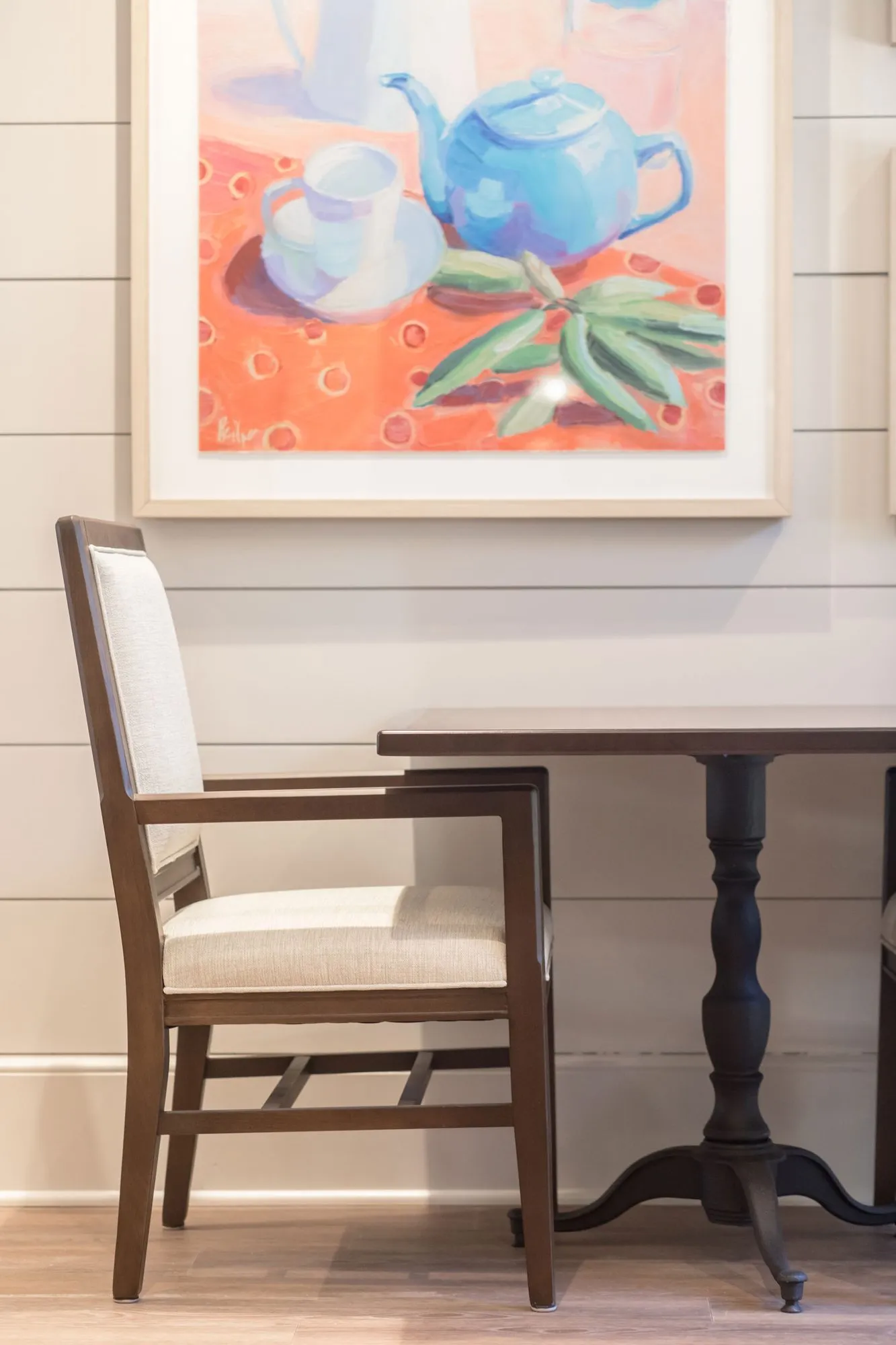 Senior living interior detail: upholstered dining chair at a pedestal table below a painting of a teapot and teacup