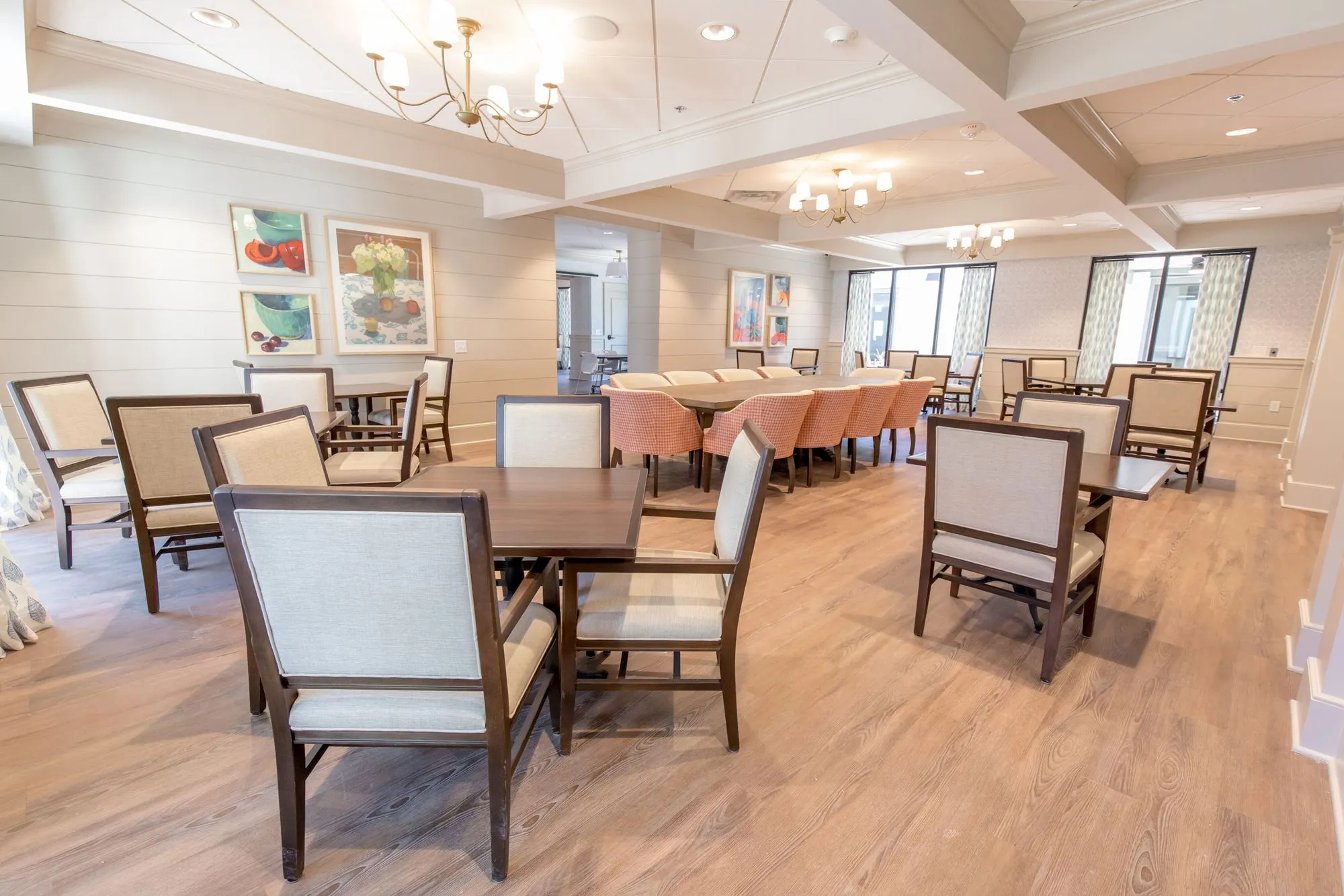 Senior living dining room with cream upholstered chairs at wood tables, brass chandeliers and colorful still life paintings