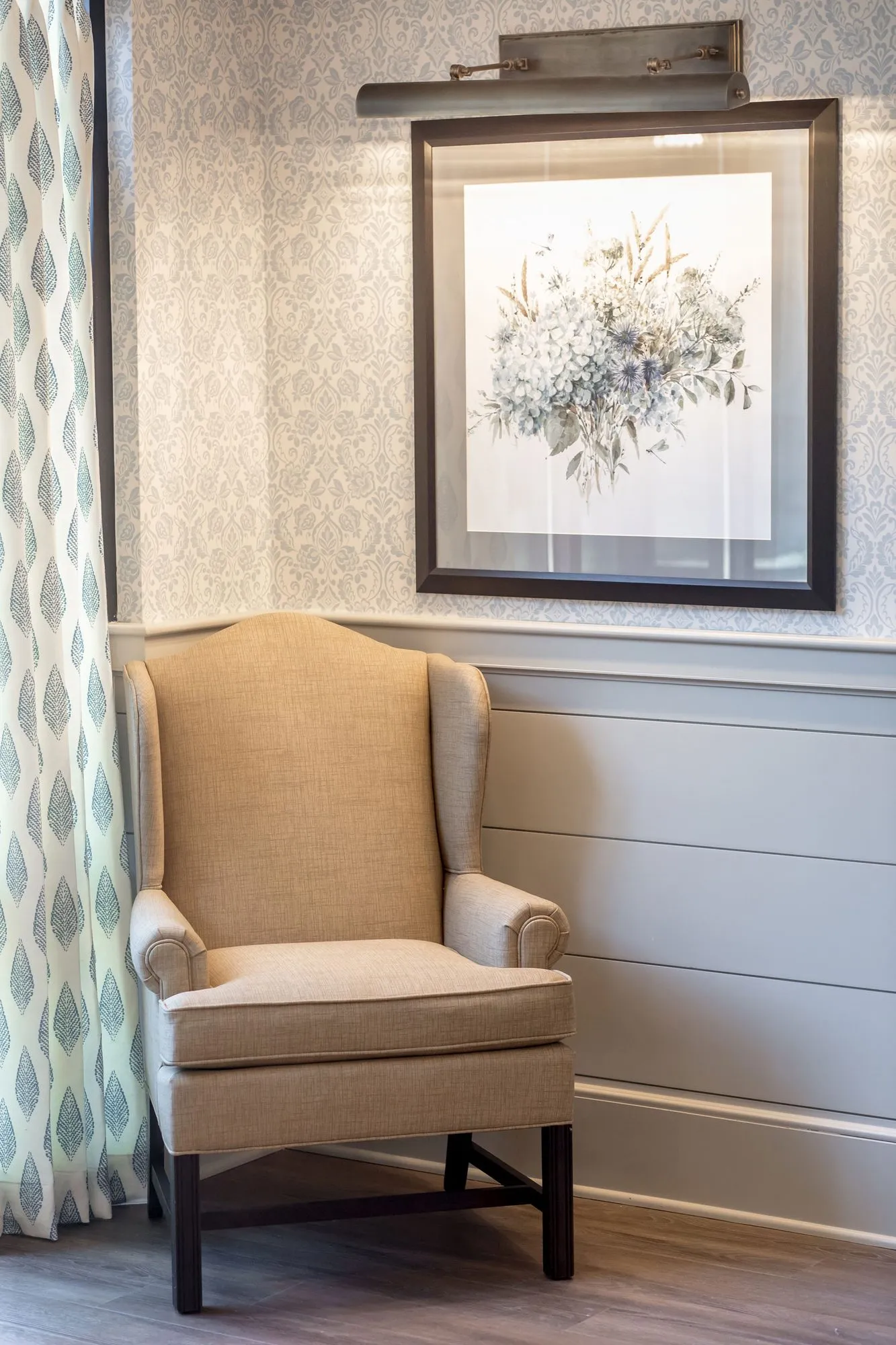 Senior living interior detail: cream wingback chair below a framed floral print and picture light, beside leaf print curtains