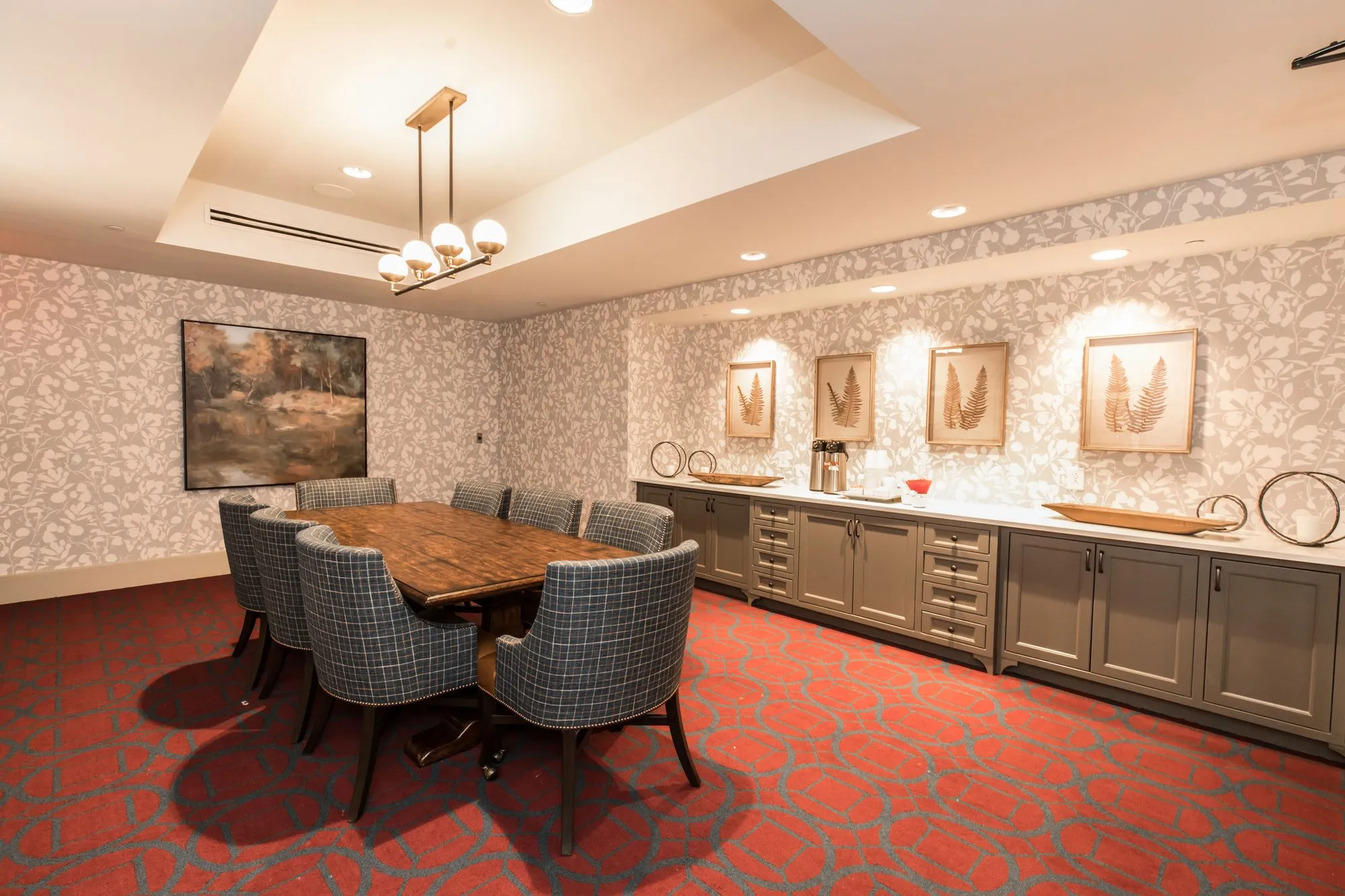 Senior living private dining room with a long rustic table, plaid chairs, red patterned carpet and botanical prints
