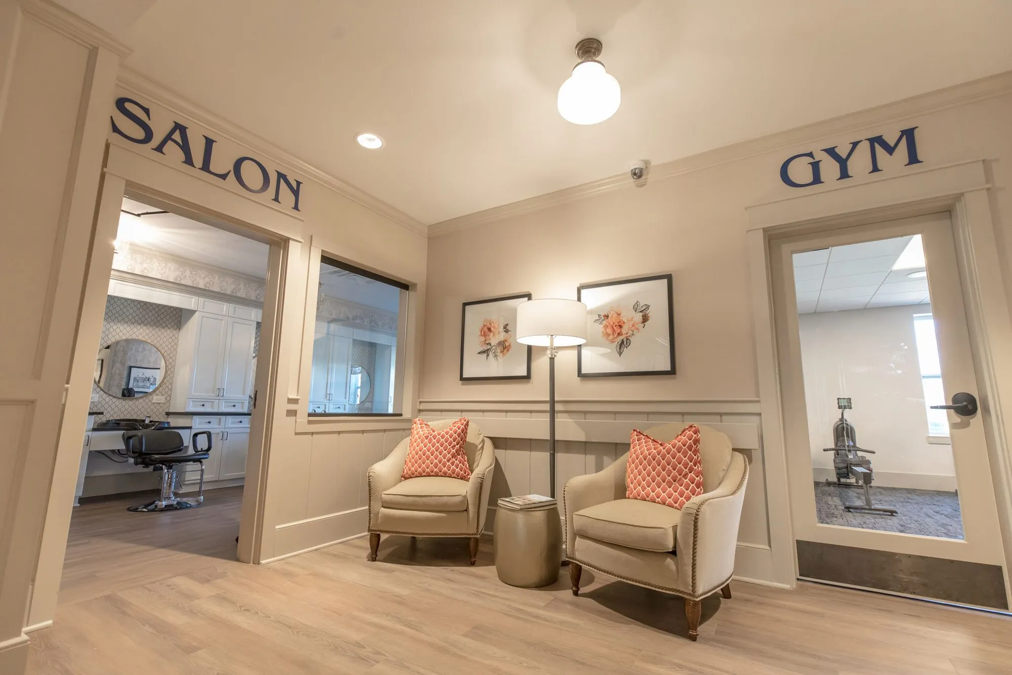 Senior living waiting area with two cream armchairs and coral pillows between doors lettered Salon and Gym