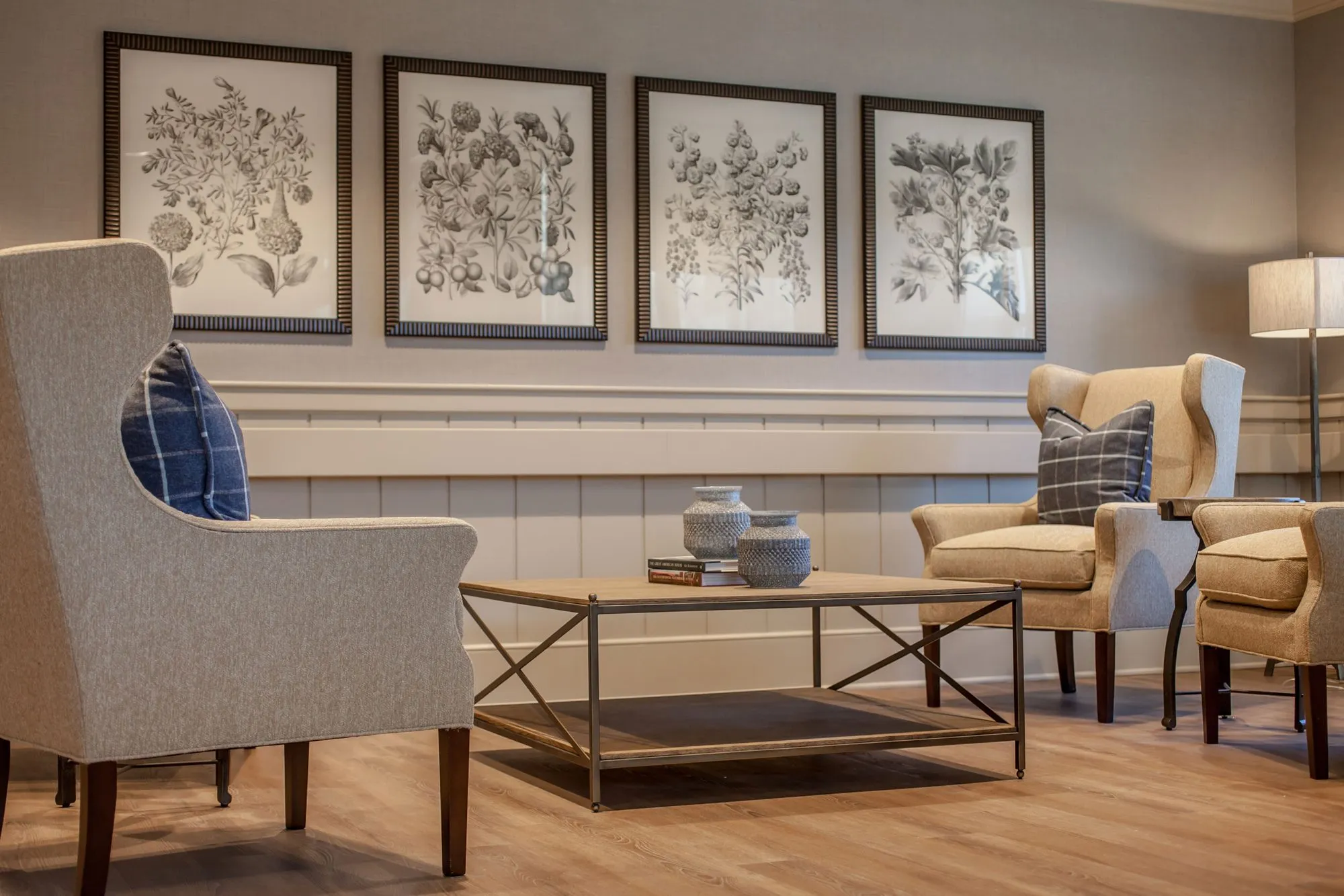 Senior living sitting area with cream wingback chairs and a metal coffee table below four framed botanical prints
