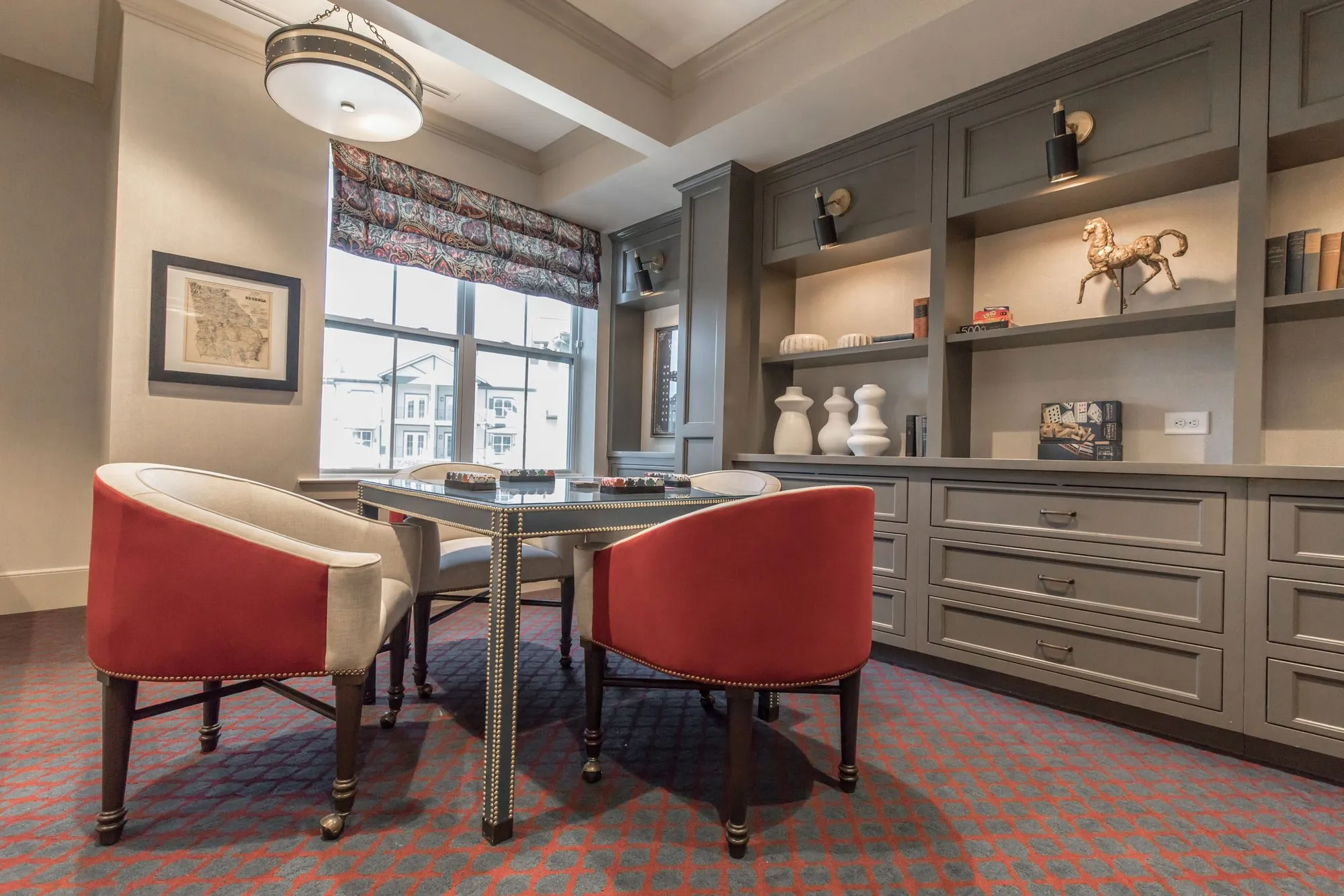 Senior living game room with a card table and red backed barrel chairs before grey built-in shelves and a patterned shade