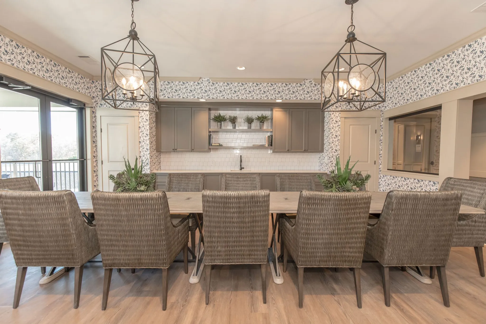 Senior living activity room with woven chairs along a long wood table under lantern pendants and floral wallcovering