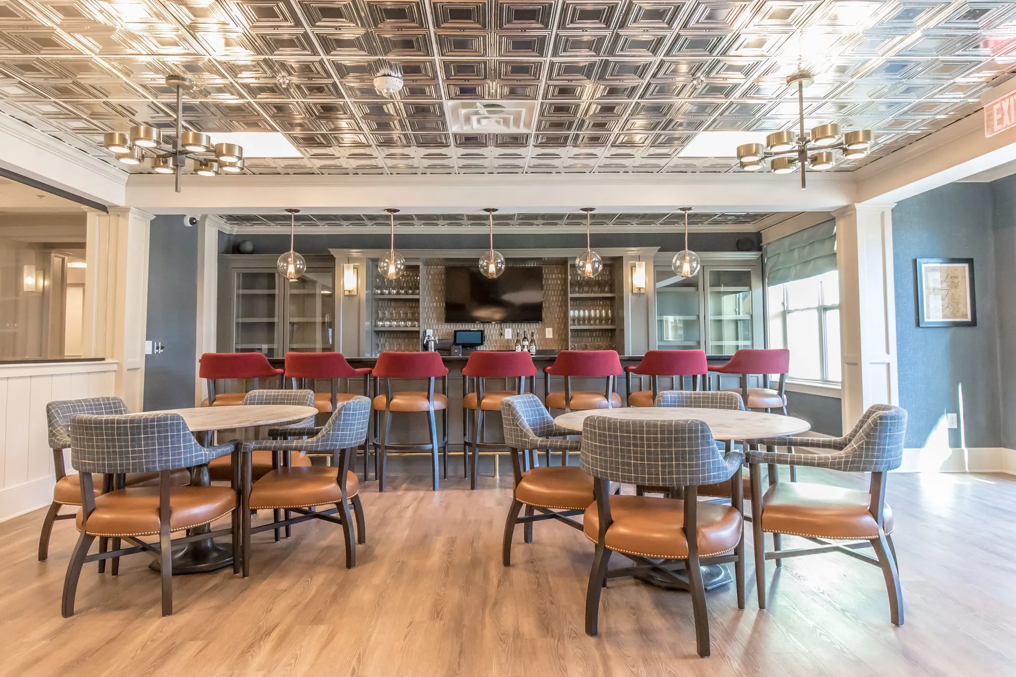 Senior living bar with red stools along the navy bar beneath a pressed tin ceiling, plaid and leather chairs at the tables
