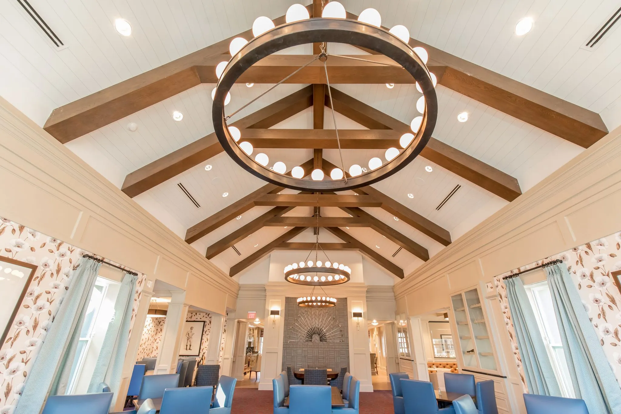 Senior living dining room under timber trusses and ring chandeliers, blue chairs and floral wallpaper toward a fireplace