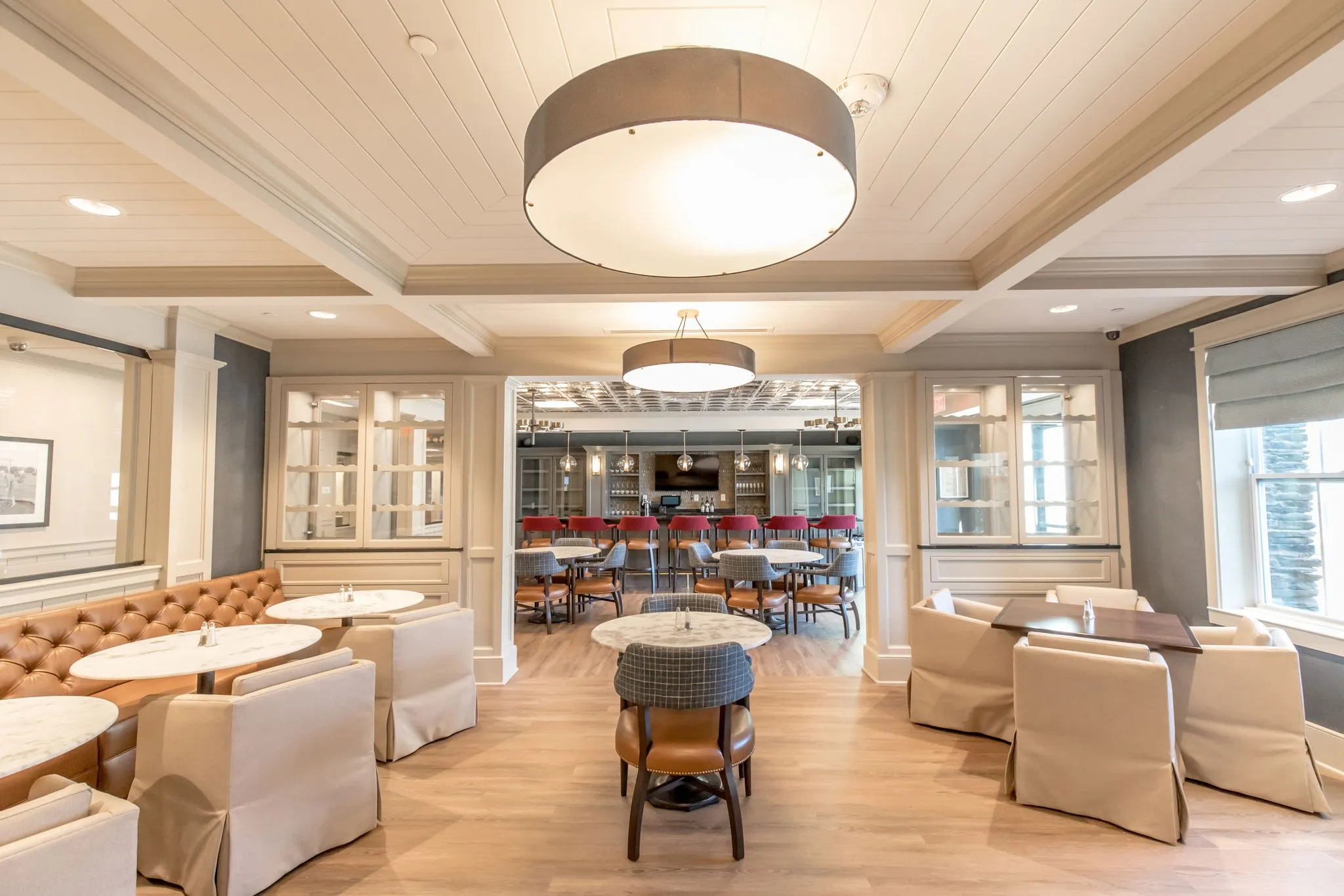 Senior living lounge with a tufted leather banquette and white granite tables under a drum pendant, the bar beyond